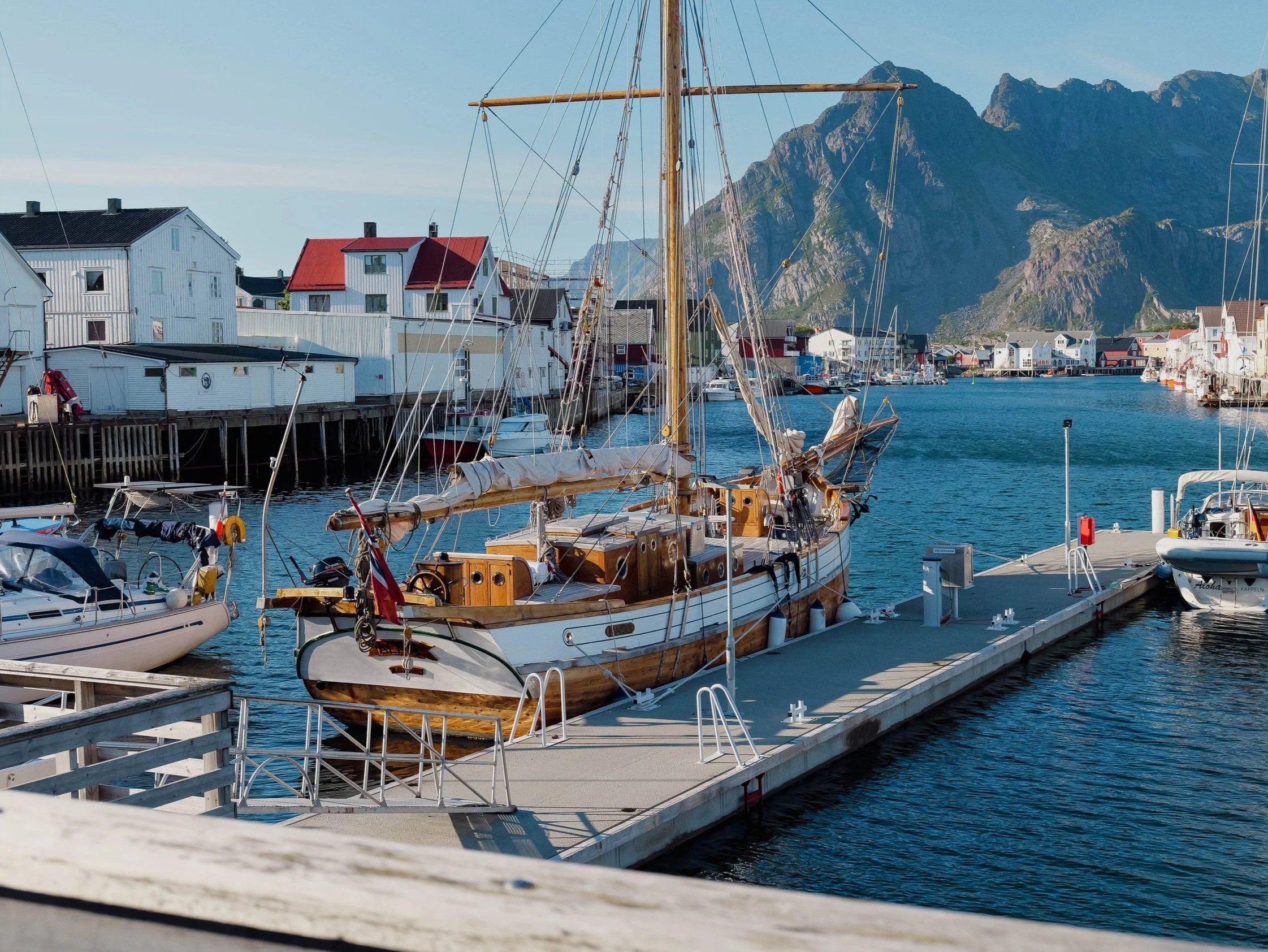 A marina with sailboats moored at a dock, colorful houses along the waterfront, and mountains in the background.