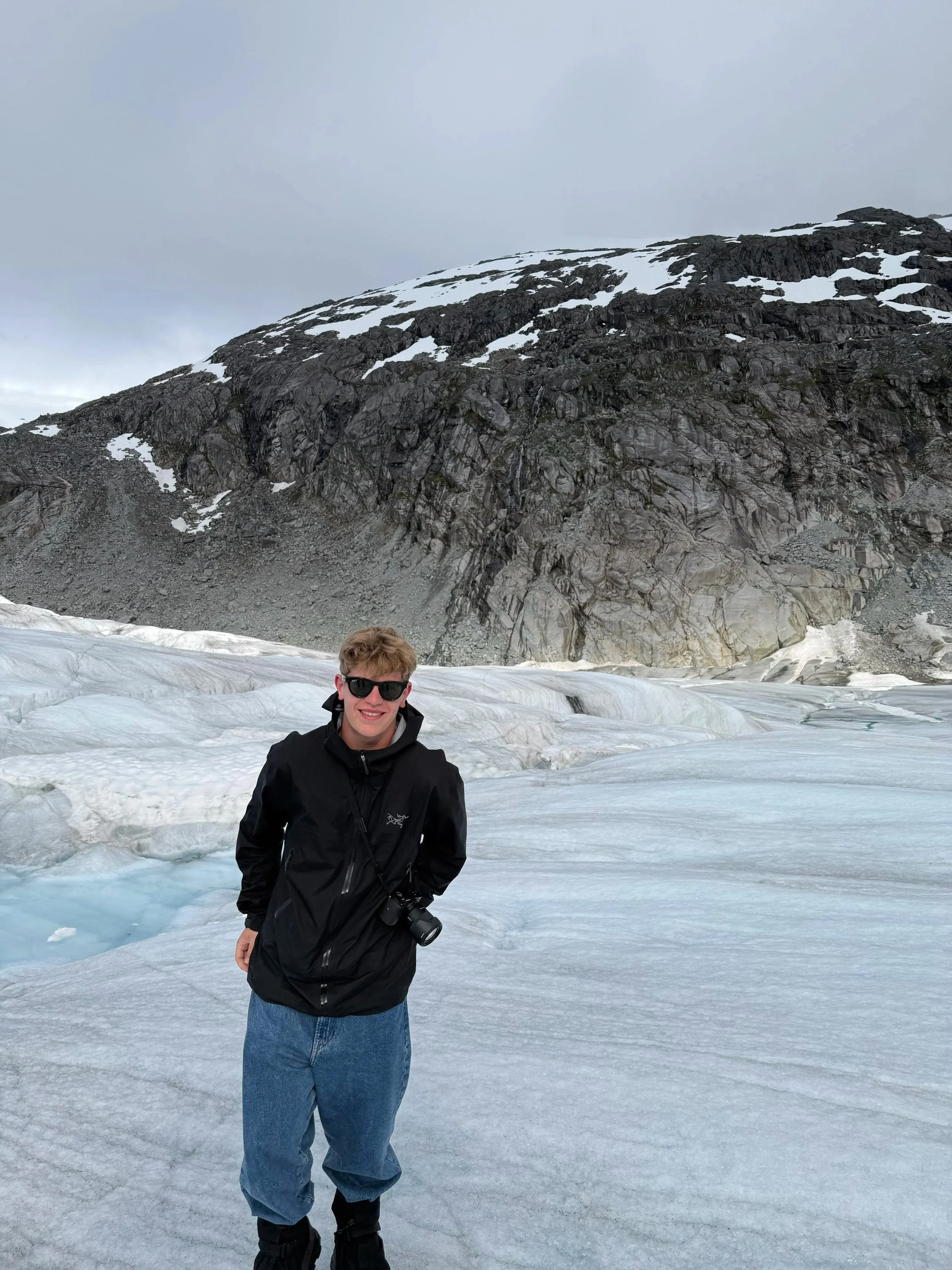 Young man wearing sunglasses and a black jacket standing on snow and ice with a mountain and glacier in the background.