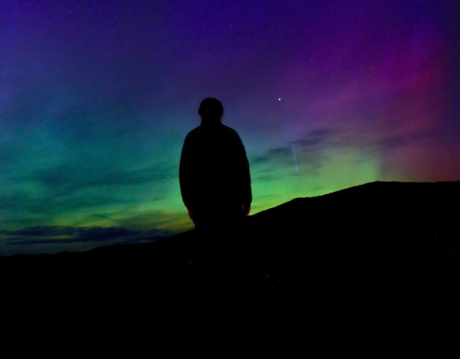 Silhouette of a person standing on a dark hilltop under a colorful night sky with aurora borealis and stars.