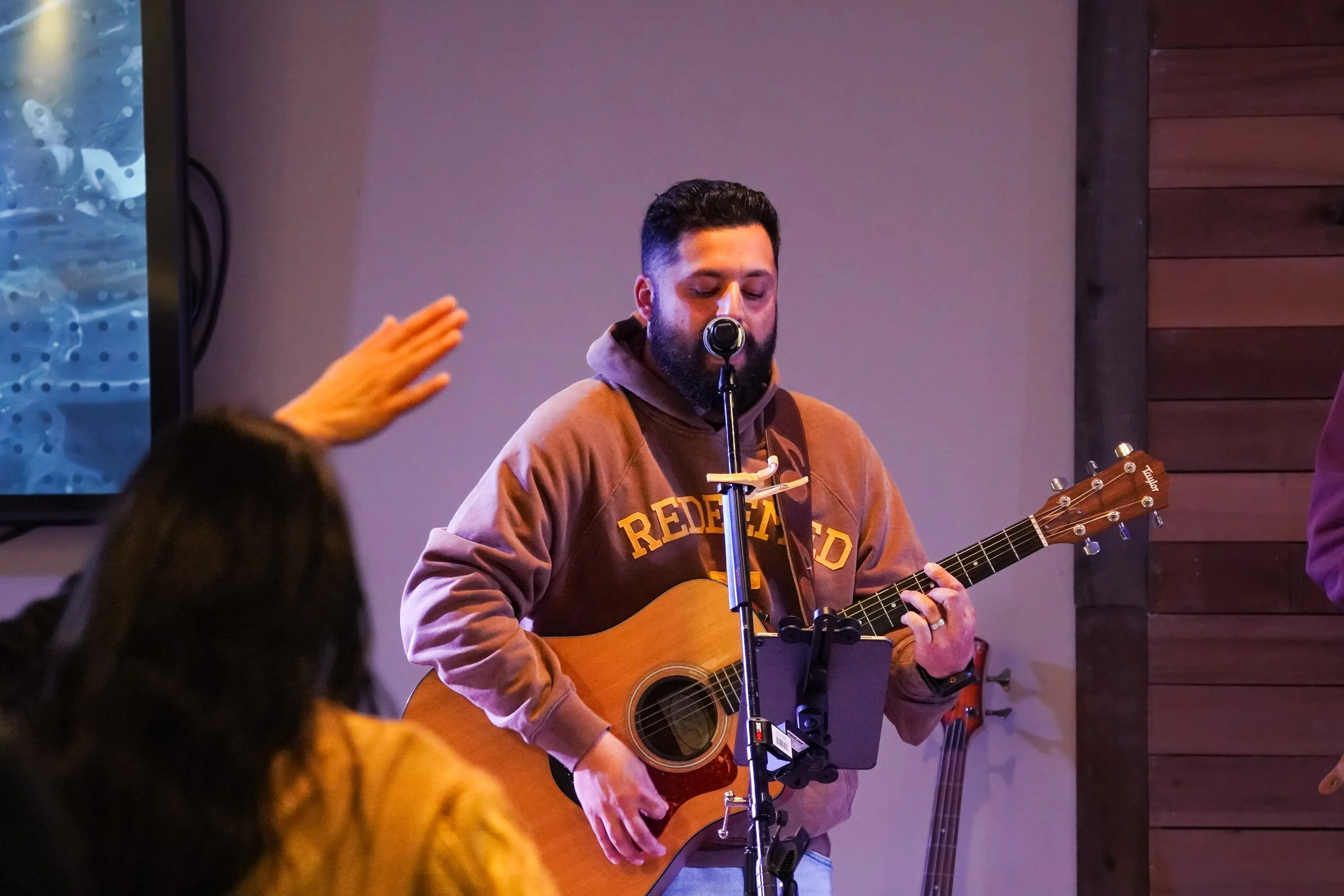 A man with a beard wearing a brown hoodie with the text 'REDEEMED' on it, playing an acoustic guitar and singing into a microphone in a dimly lit indoor setting. An audience member with dark hair and a yellow jacket is raising their hand in front of him.