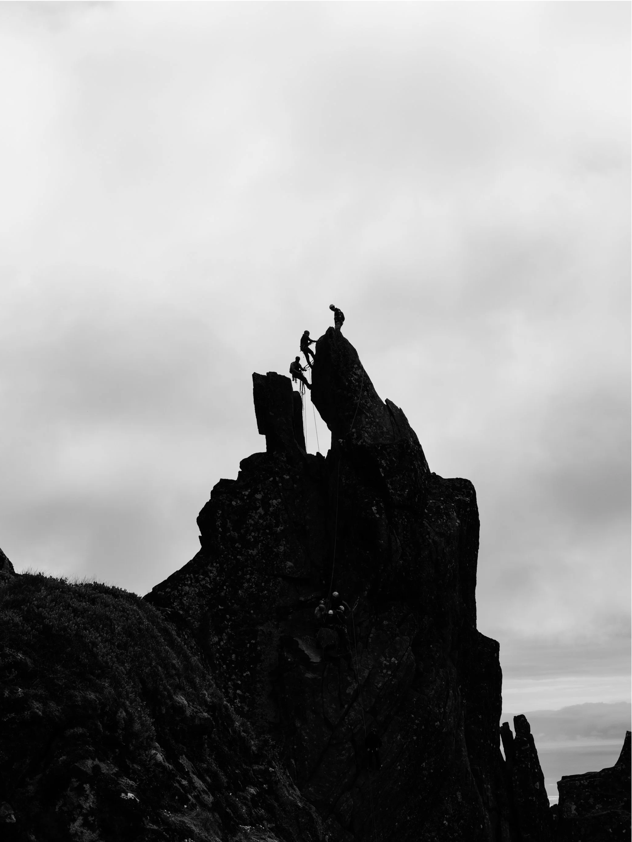 Silhouetted rock climbers ascending a steep rock formation against a cloudy sky.