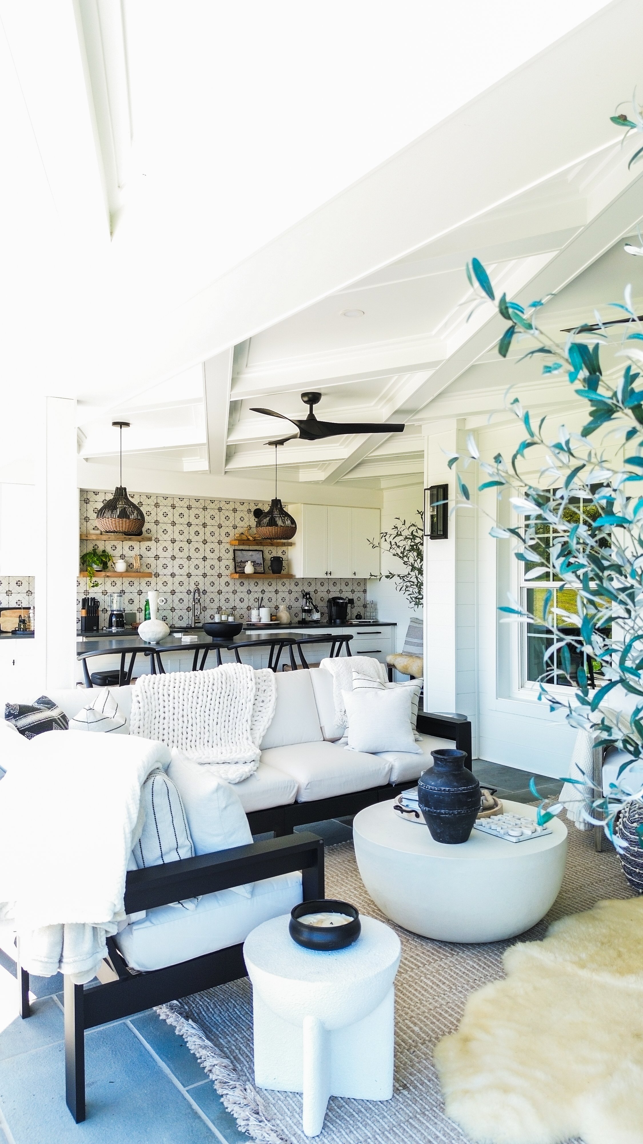 Living room with white sofa, black coffee table, and decorative vases; open kitchen with patterned backsplash, hanging pendant lights, and barstools in the background.