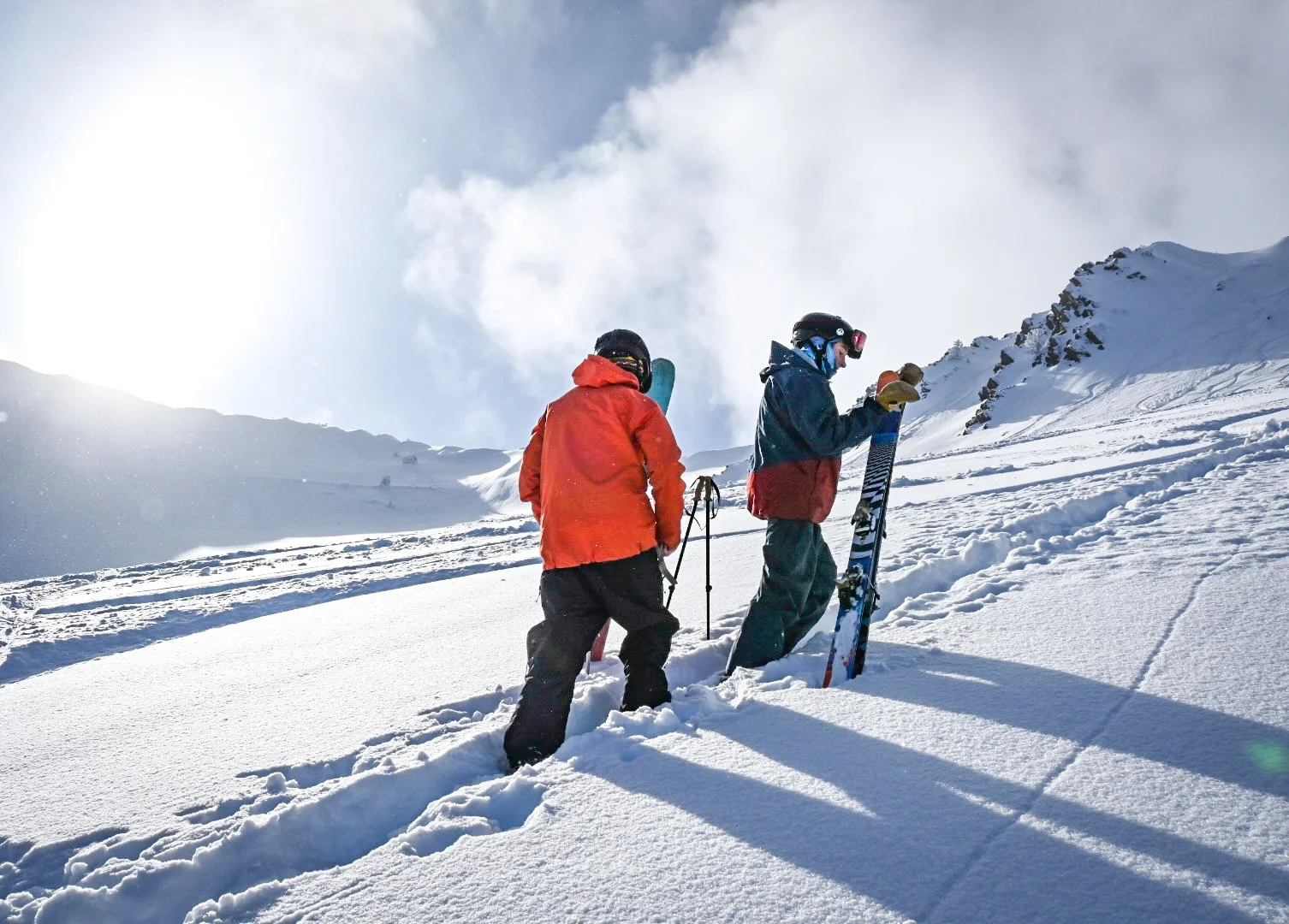 Two people in winter gear walking up a snowy mountain slope, one holding ski poles and the other holding a pair of skis, with snow-covered mountain peaks in the background under a partly cloudy sky.