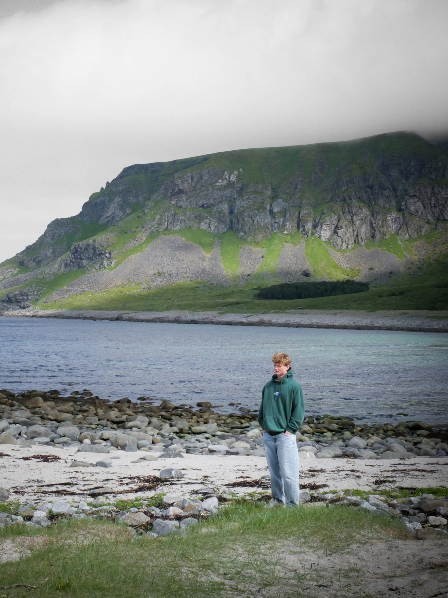 A young man with blond hair in a green hoodie and light blue jeans standing on a grassy and rocky beach with a mountain, a body of water, and an overcast sky in the background.