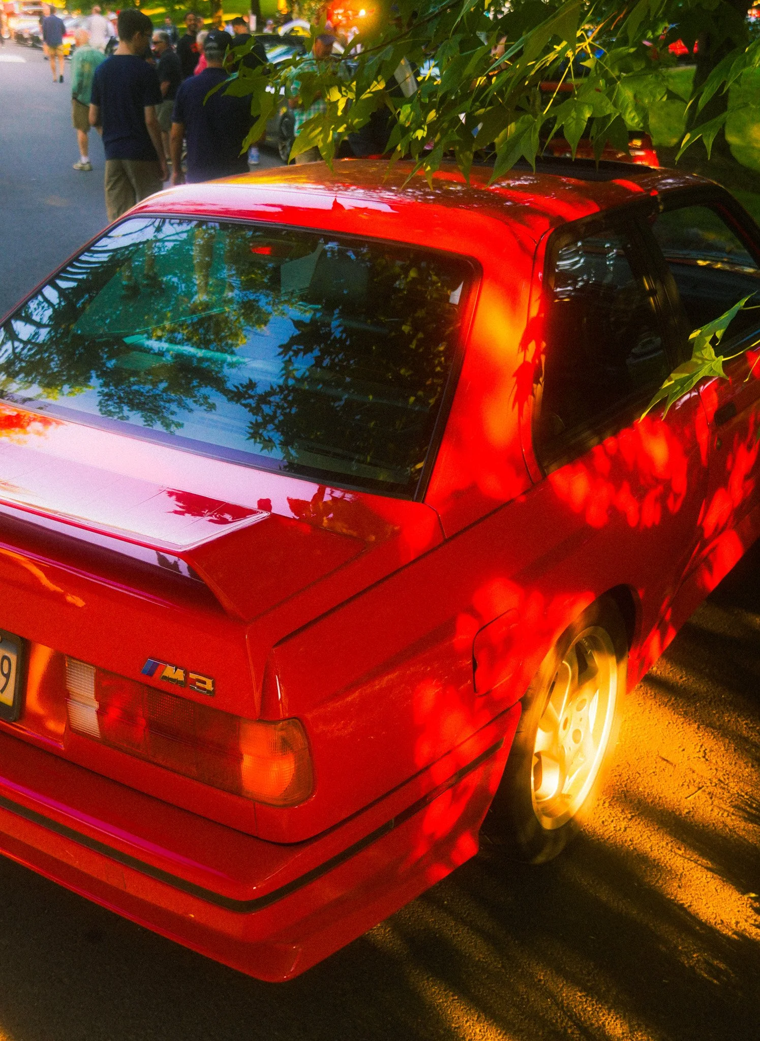 A red BMW M3 parked under trees, with sunlight filtering through the leaves and casting shadows on the car. People are gathered in the background.