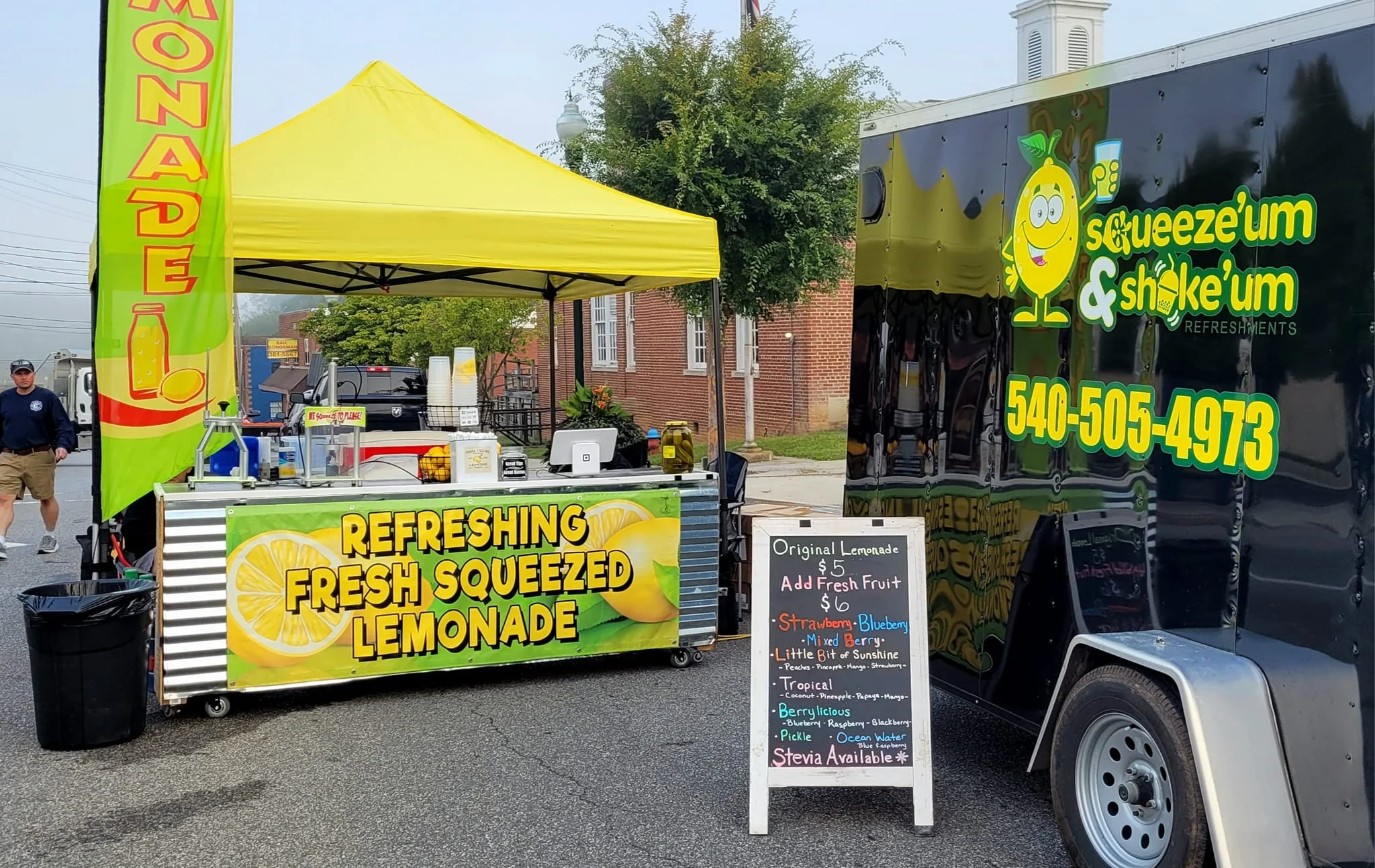 A food stand selling refreshing lemonades at an outdoor event, featuring a bright yellow tent, a green banner, and a black trailer with the stand's name and contact number. The stand offers fresh squeezed lemonades with various flavors listed on a chalkboard sign.