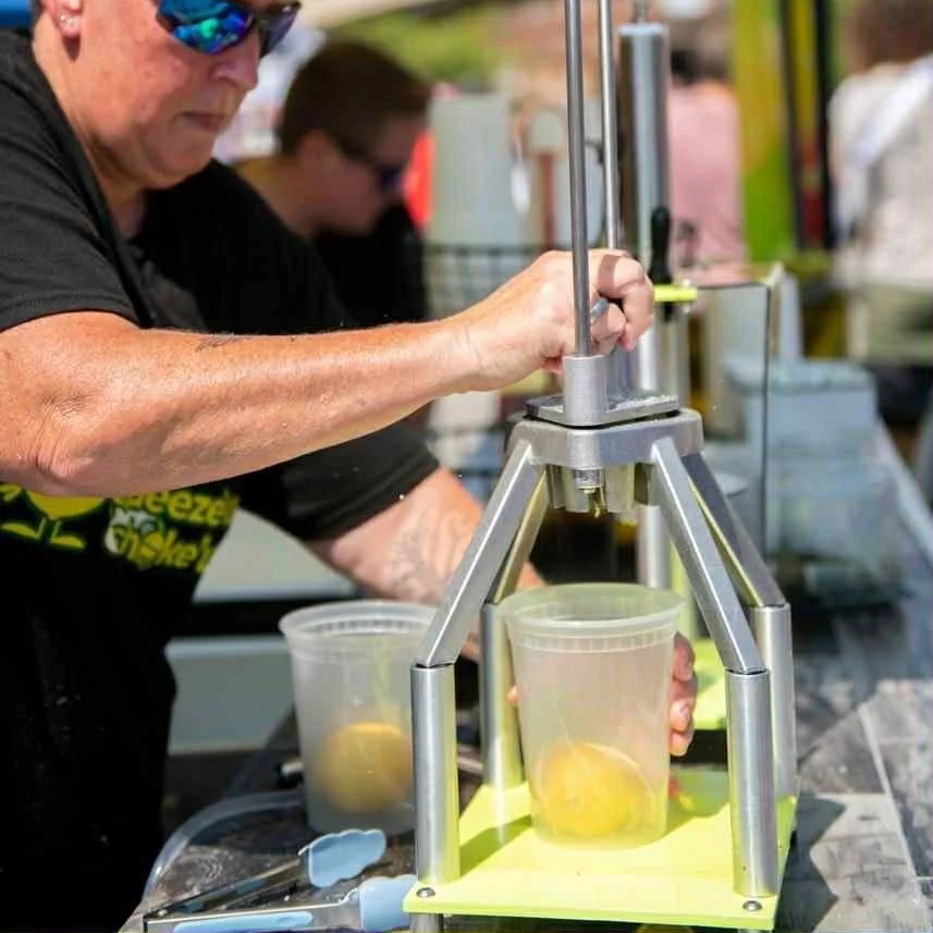 Person squeezing lemonade from a lemon press at an outdoor booth, with a plastic cup catching the juice, other lemons and tools on the table.