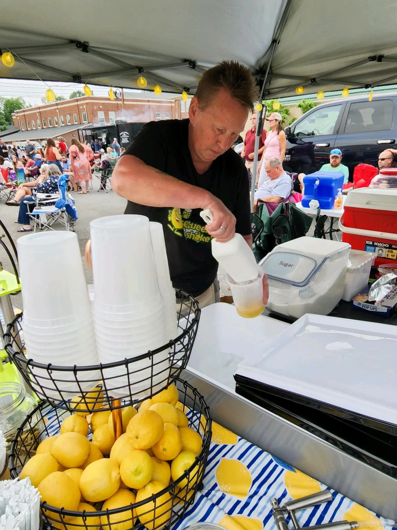 A man in a black T-shirt preparing a beverage at an outdoor event tent, with a basket of yellow lemons, disposable cups, and supplies on the table. There are people and vehicles in the background.
