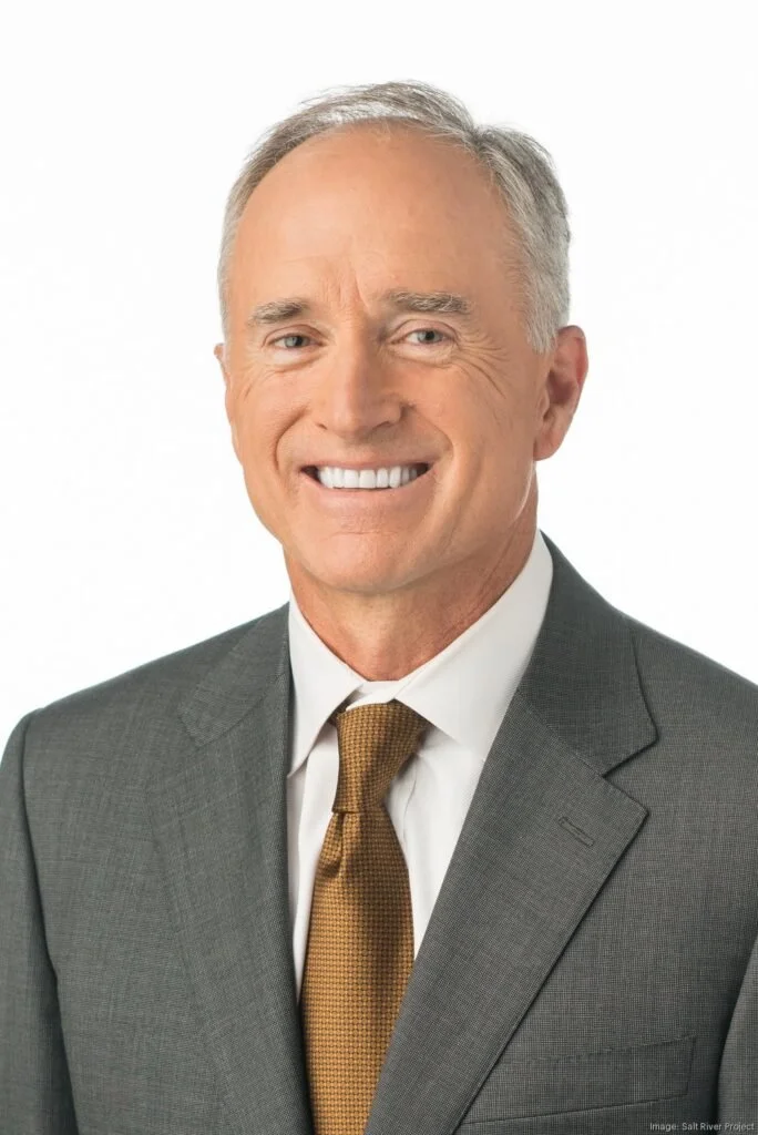 Headshot of smiling middle-aged man with gray hair, wearing a gray suit, white shirt, and brown tie against a white background.