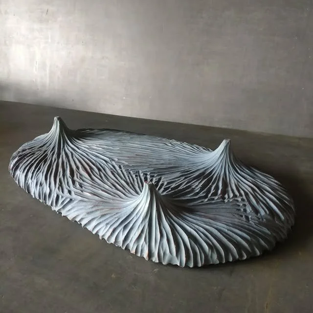 Skulptur in Form eines spiralförmigen, wellenartigen Objekts mit rillenförmiger Oberfläche auf einem Boden in einem Raum mit grauer Wand.