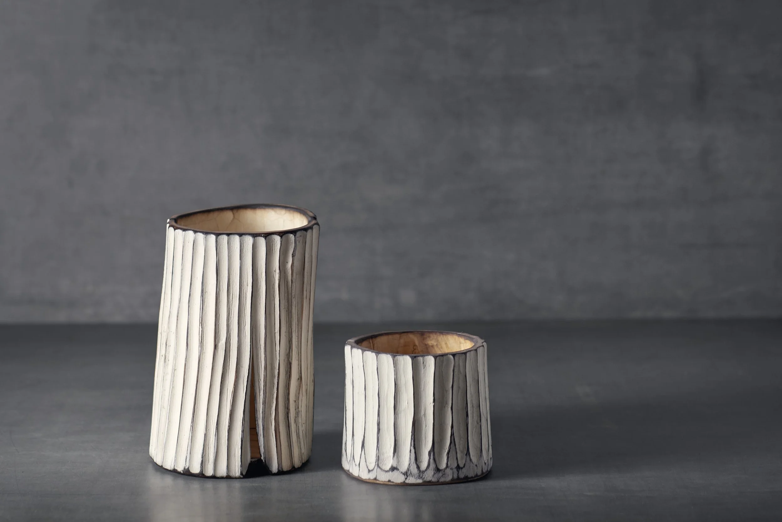 Zwei Kerzenhalter aus geflochtenem Holz auf grauem Hintergrund, unterschiedlich hoch und breit.
