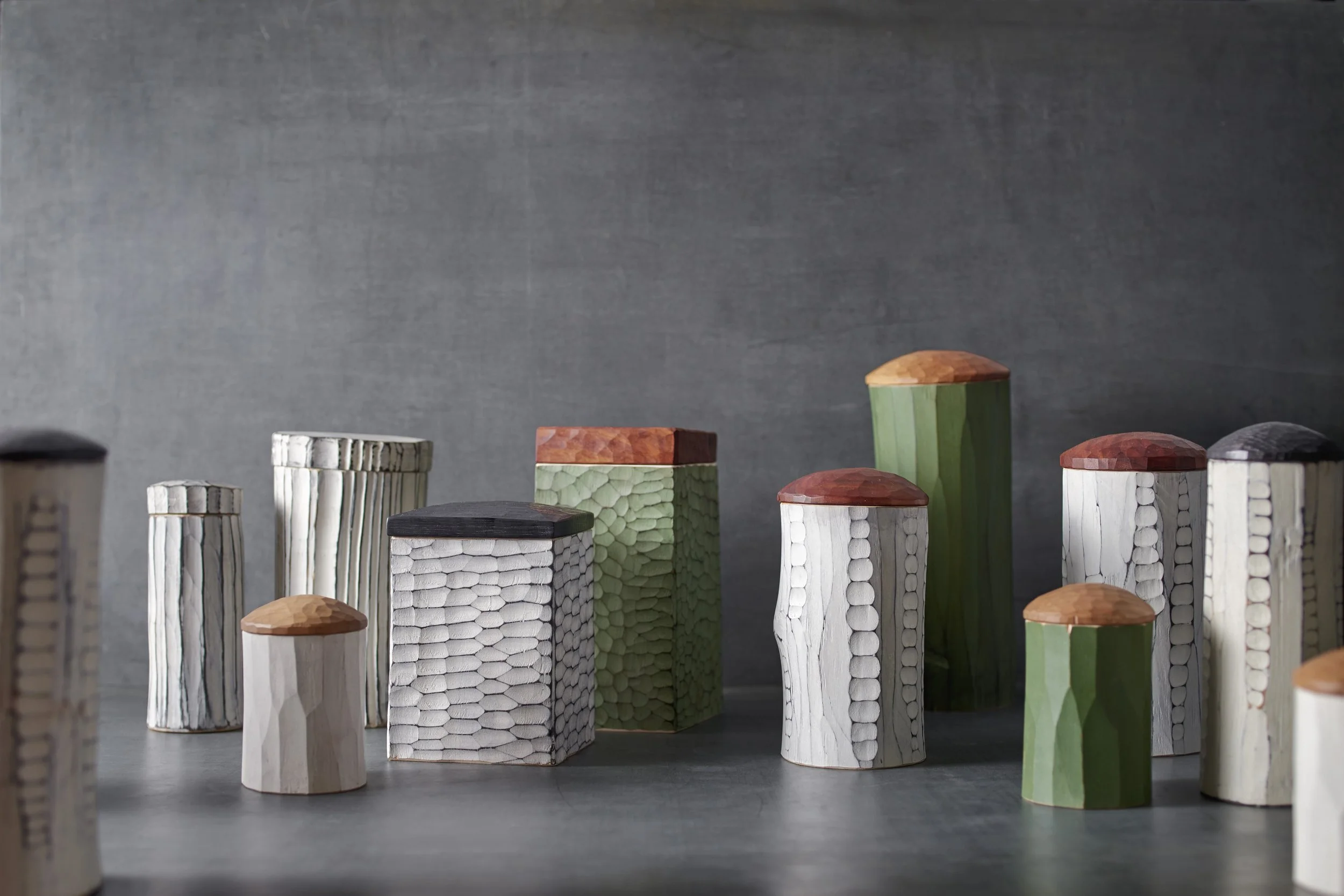 Mehrere geschnitzte Holzdosen oder Urnen. Die Oberfläche ist mit geschnitzter Textur und Ornamentik versehen und weiß und grün bemalt. Die eckigen und runden Gefße sind mit abgerundeten und eckigen Deckeln aus verschiedenen Hölzern kombiniert.