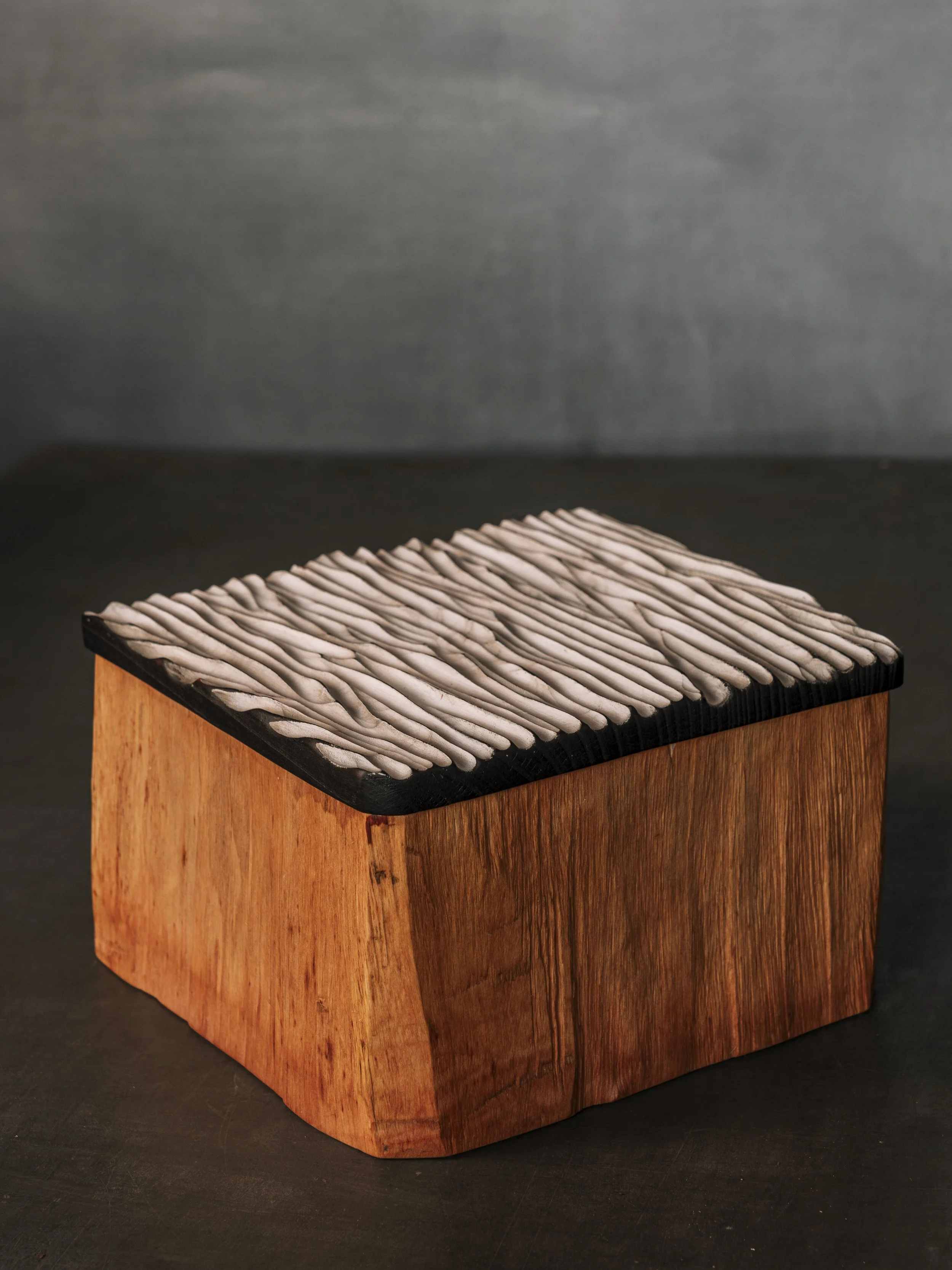 Eckiges Holzgefäß oder Urne . Die Wandung wurde eckig gespalten aus einem massiven Stück Baumstamm. Die Wandung Naturfarben, der Deckel mit geriffelter Struktur in schwarz und weiß.