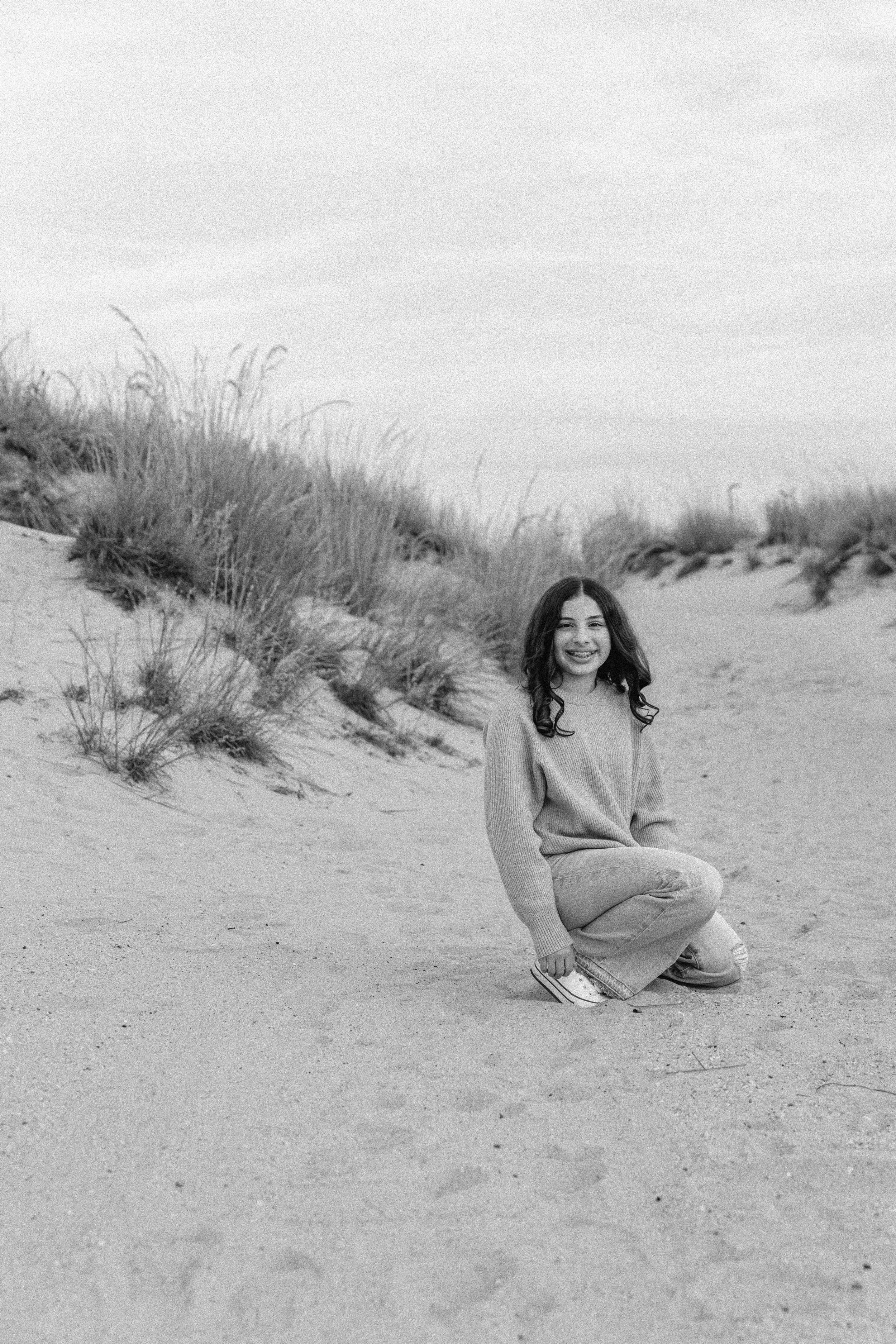 A smiling young woman with dark wavy hair crouches on a sandy beach with dunes and grass in the background, captured in black and white.