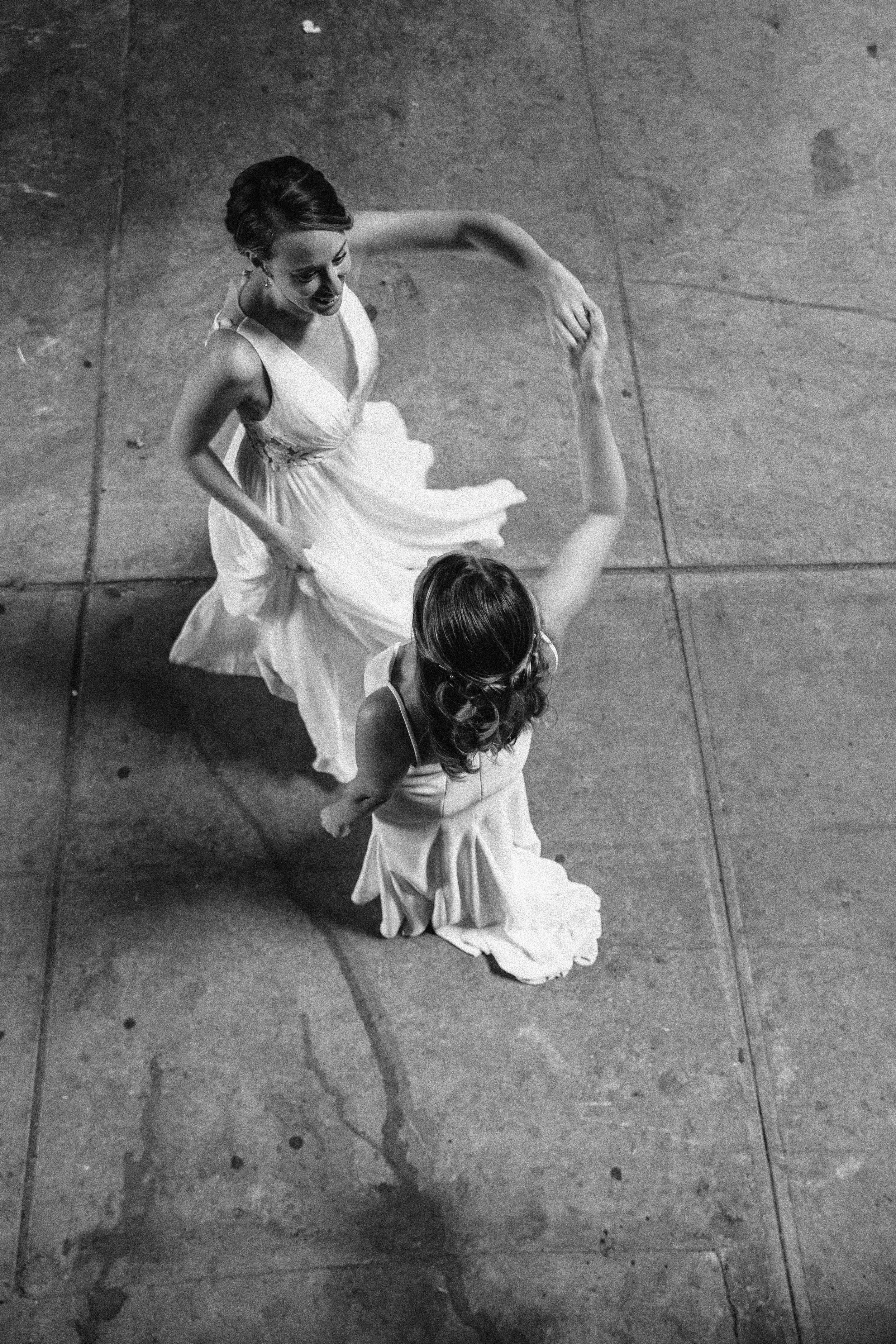 Two women in flowing dresses dance on a concrete floor, holding hands and forming a circle. Documentary wedding photography in NYC