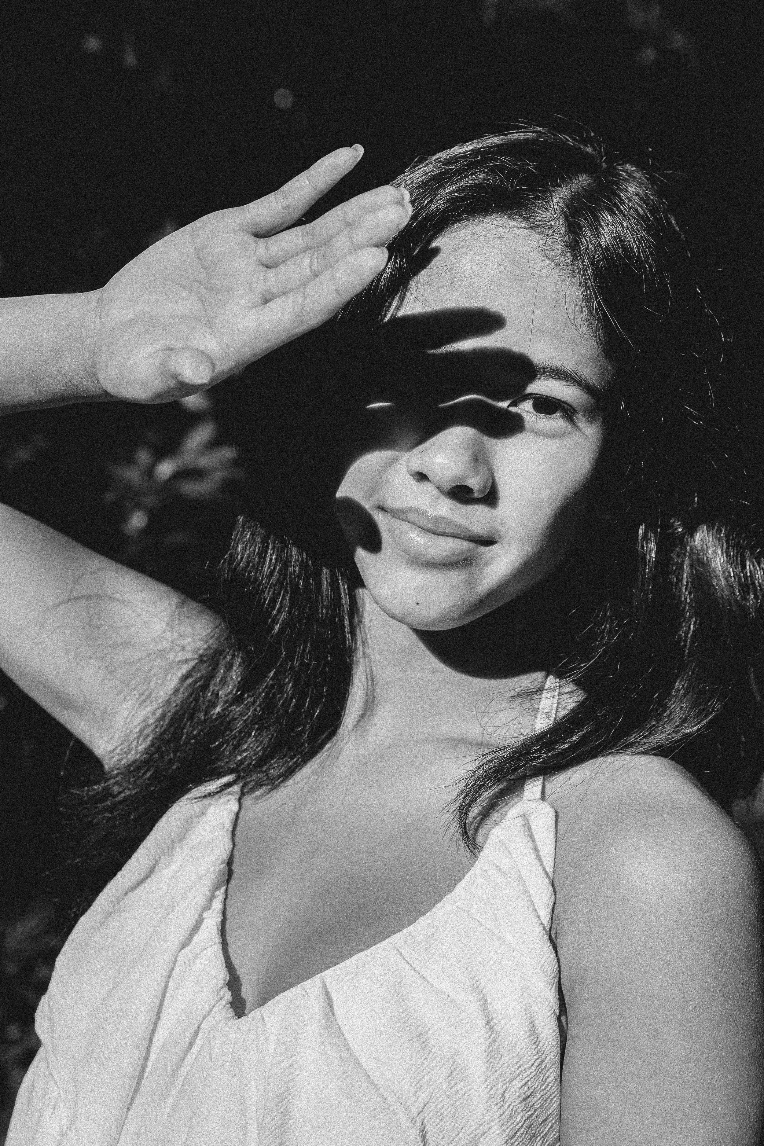 Black and white photo of a woman with long hair, partially covering her face with her hand, casting a shadow over one eye, with a slight smile.