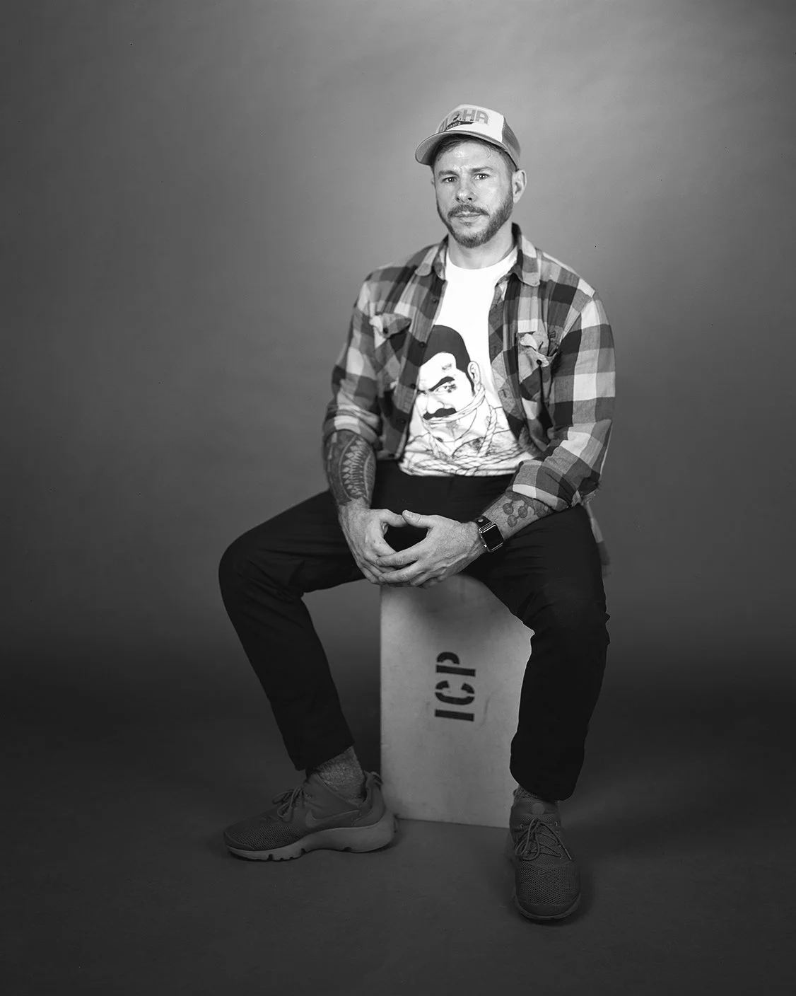 A man with tattoos, a beard, and a cap sitting on a box labeled 'IC9'. He is wearing a graphic t-shirt, checkered shirt, black pants, and sneakers, posing against a plain background.