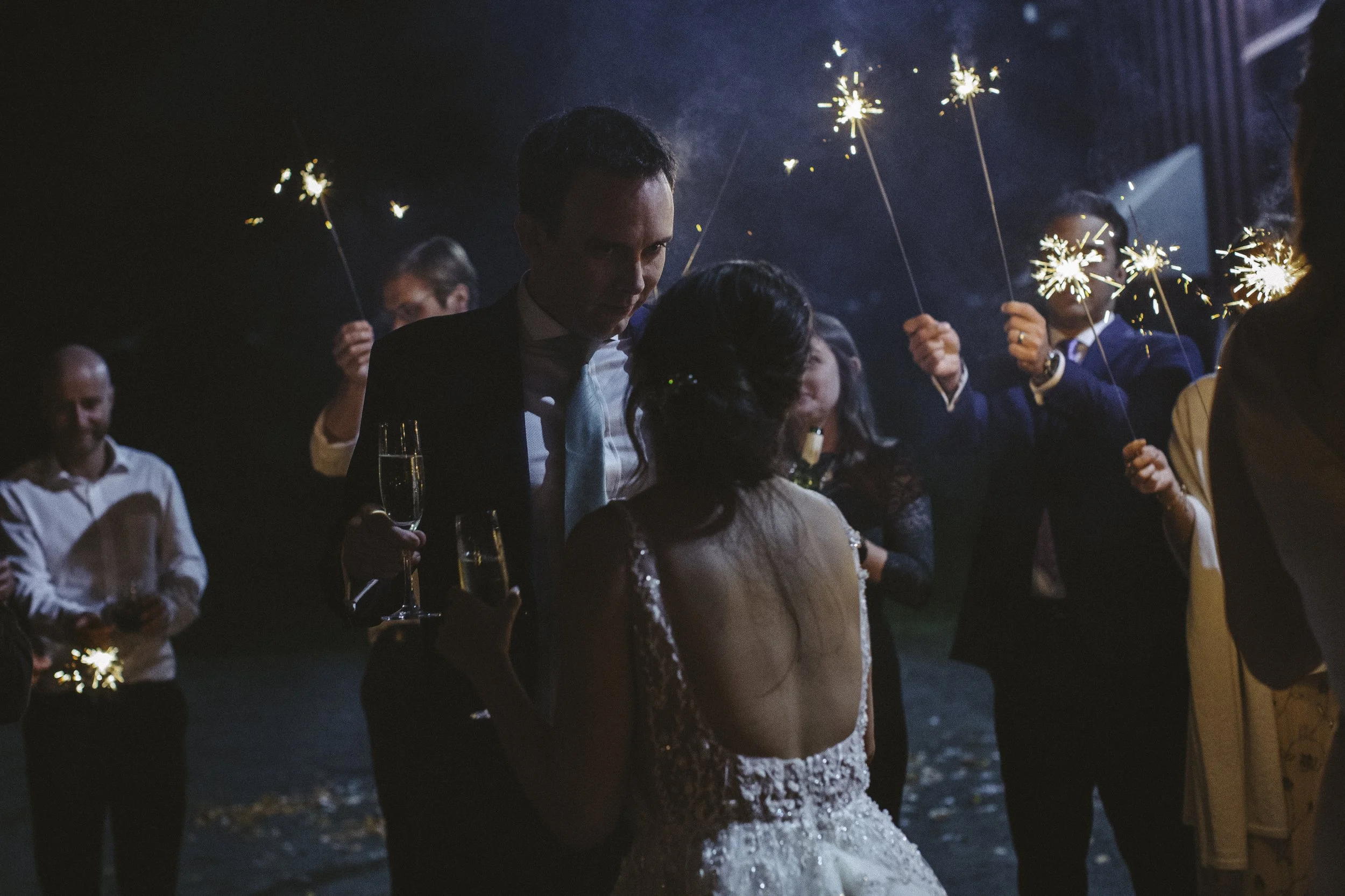 Couple at a wedding celebration with sparklers, champagne glasses, and guests in the background at night.