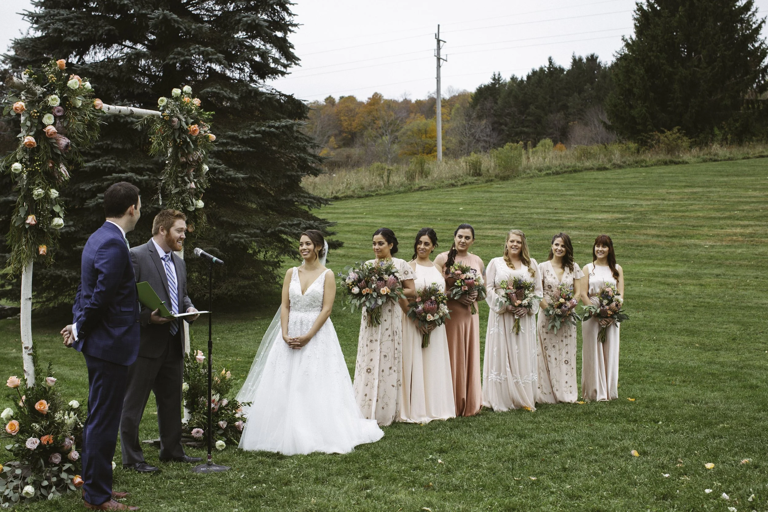 Wedding ceremony outdoors with bride, groom, and bridesmaids under a floral arch.