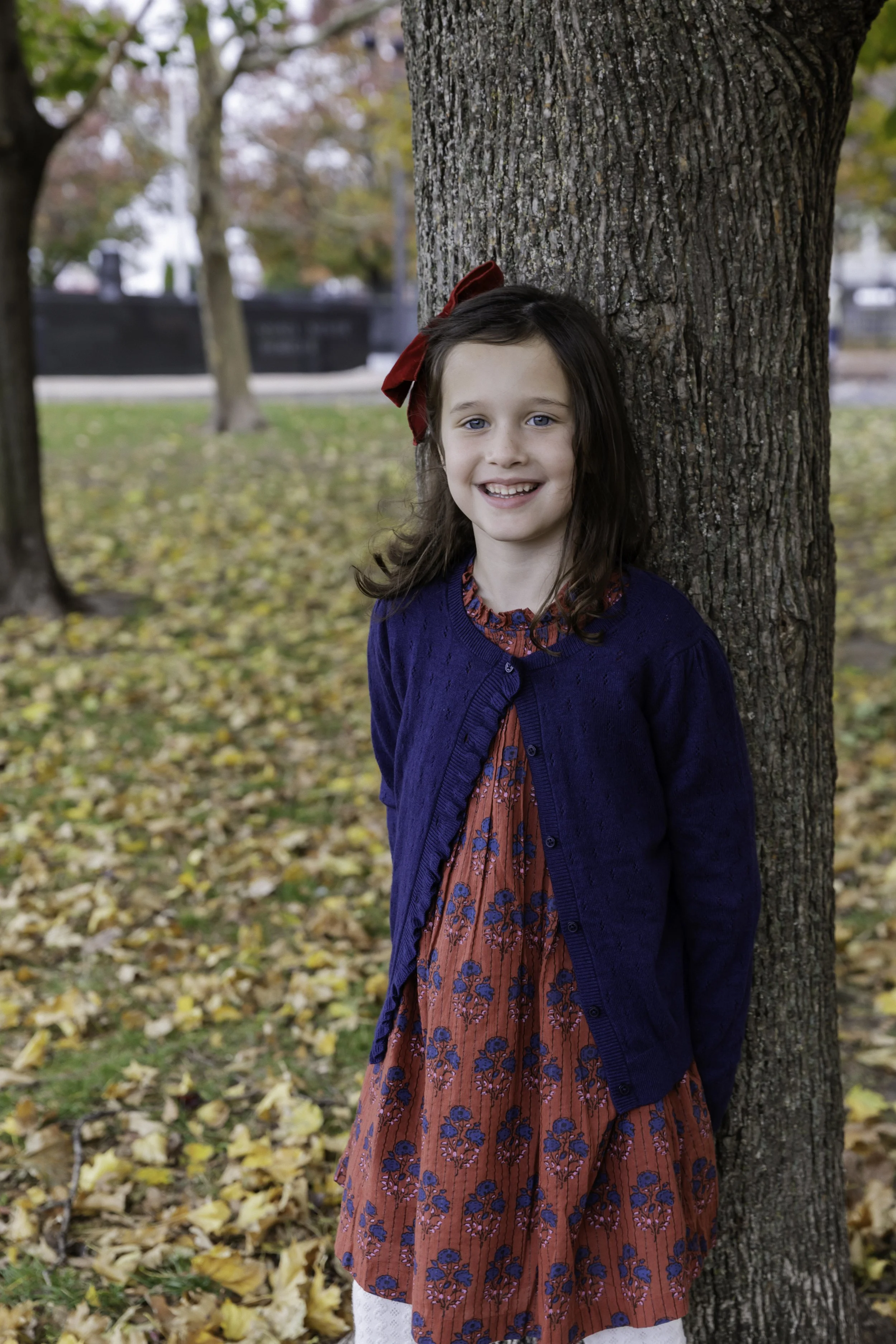 A young girl with brown hair, blue eyes, and a red bow in her hair, standing next to a tree outdoors during fall. She is wearing a blue cardigan over a red floral dress and smiling at the camera.