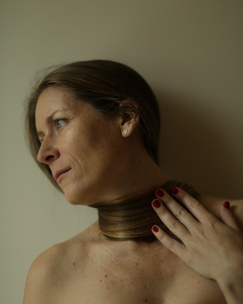 A woman with short brown hair and a gold heart-shaped earring is holding her neck with her right hand, which has red painted nails. She appears to be looking to the left with a serious expression.