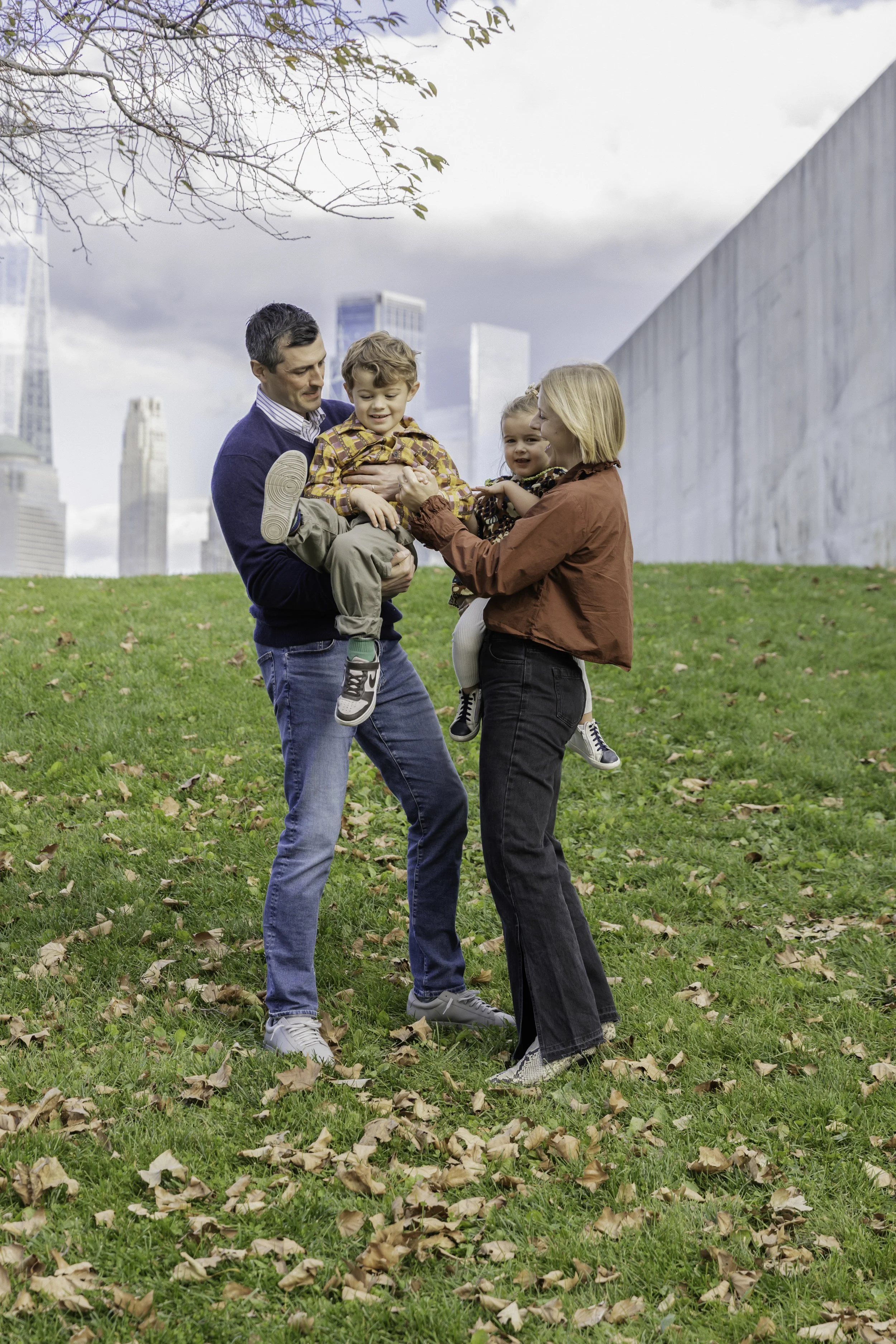 A family of four playing outdoors with tall city buildings in the background. Outdoor family portrait session in NYC, candid and natural photography