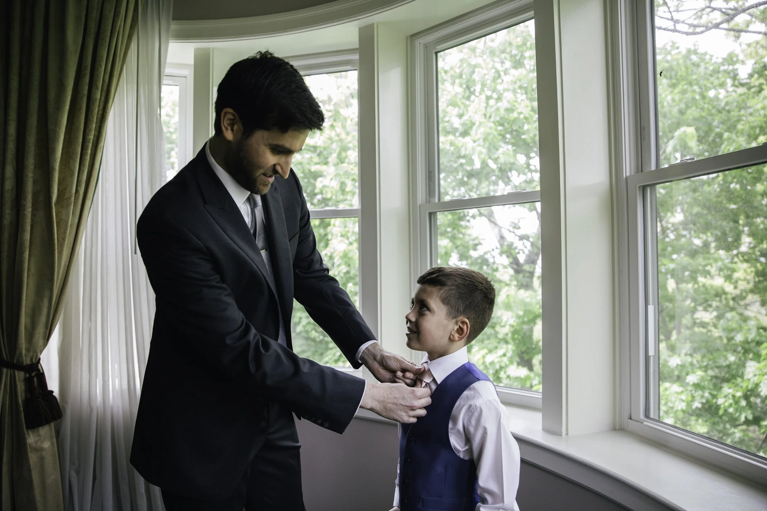 A man in a suit helps a young boy adjust his tie in front of a large window with trees outside. Documentary wedding photography in NYC