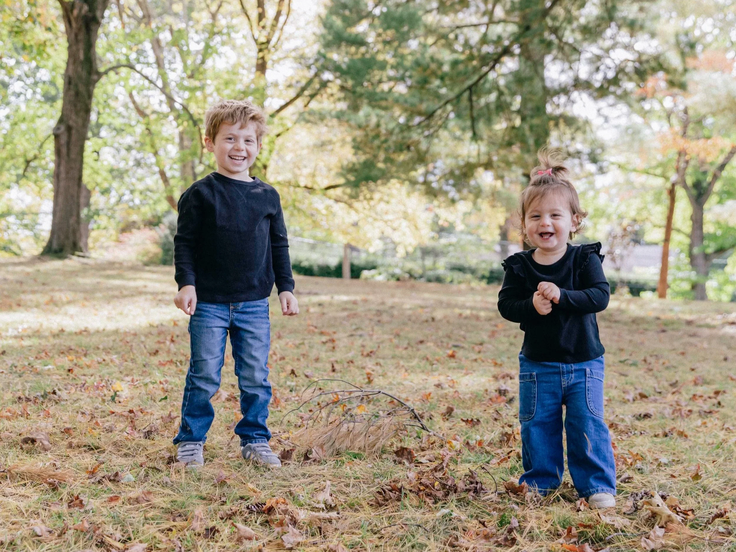 Two young children, a boy and a girl, smiling and playing outdoors in a park with autumn leaves on the ground and trees with colorful foliage in the background.