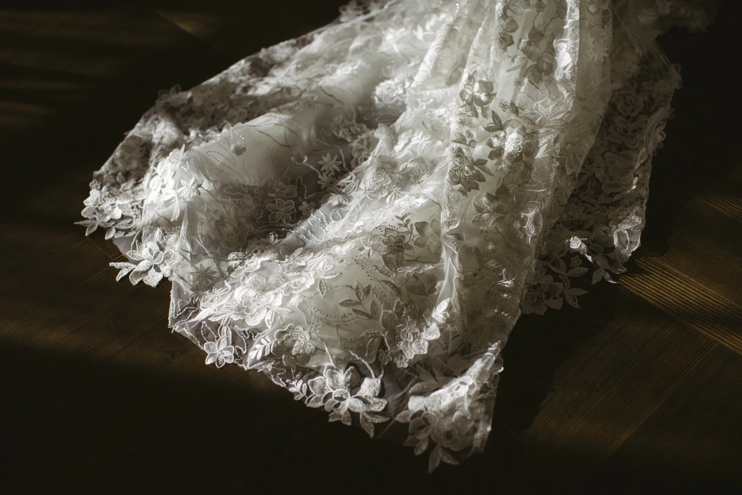 A lace wedding veil with floral embroidery, laid on a dark wooden floor with sunlight casting shadows.