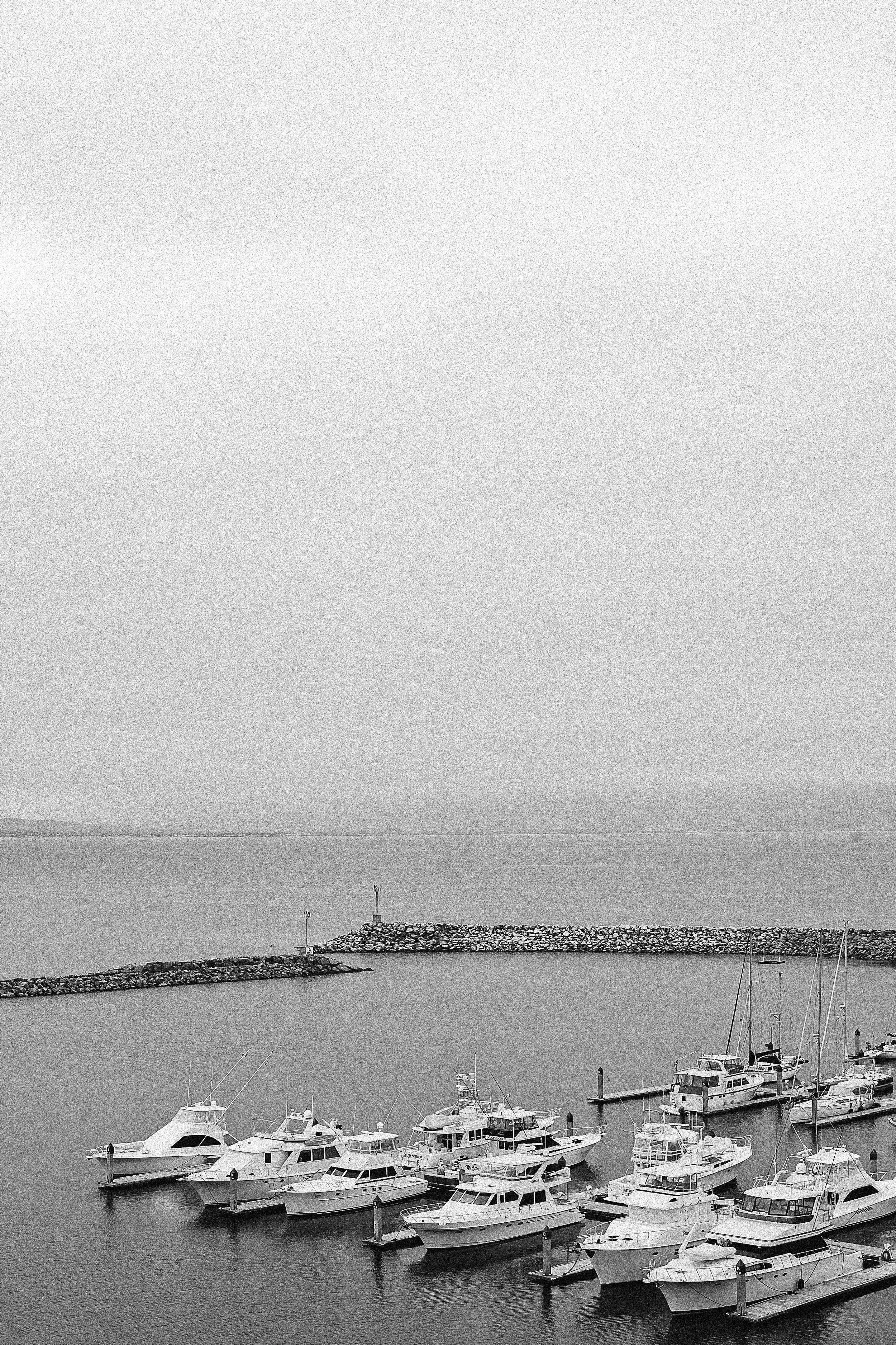 A black and white photo of yachts docked in a marina with calm water, a breakwater made of rocks, and an open sky in the background.