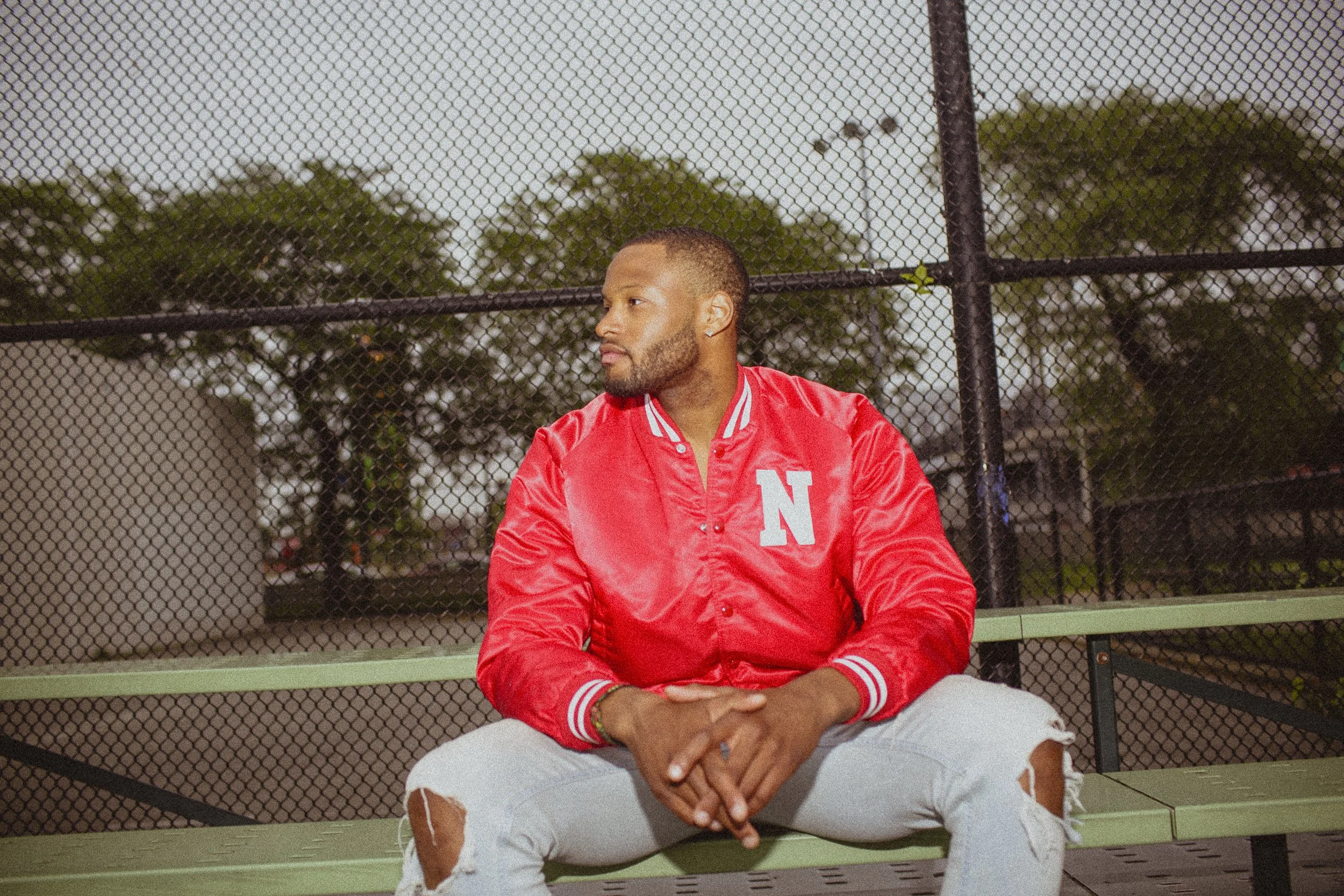A man wearing a red varsity jacket with a large 'N' on the chest, ripped light jeans, sits on a green bench behind a chain-link fence, looking to his left, outdoors in the evening.
