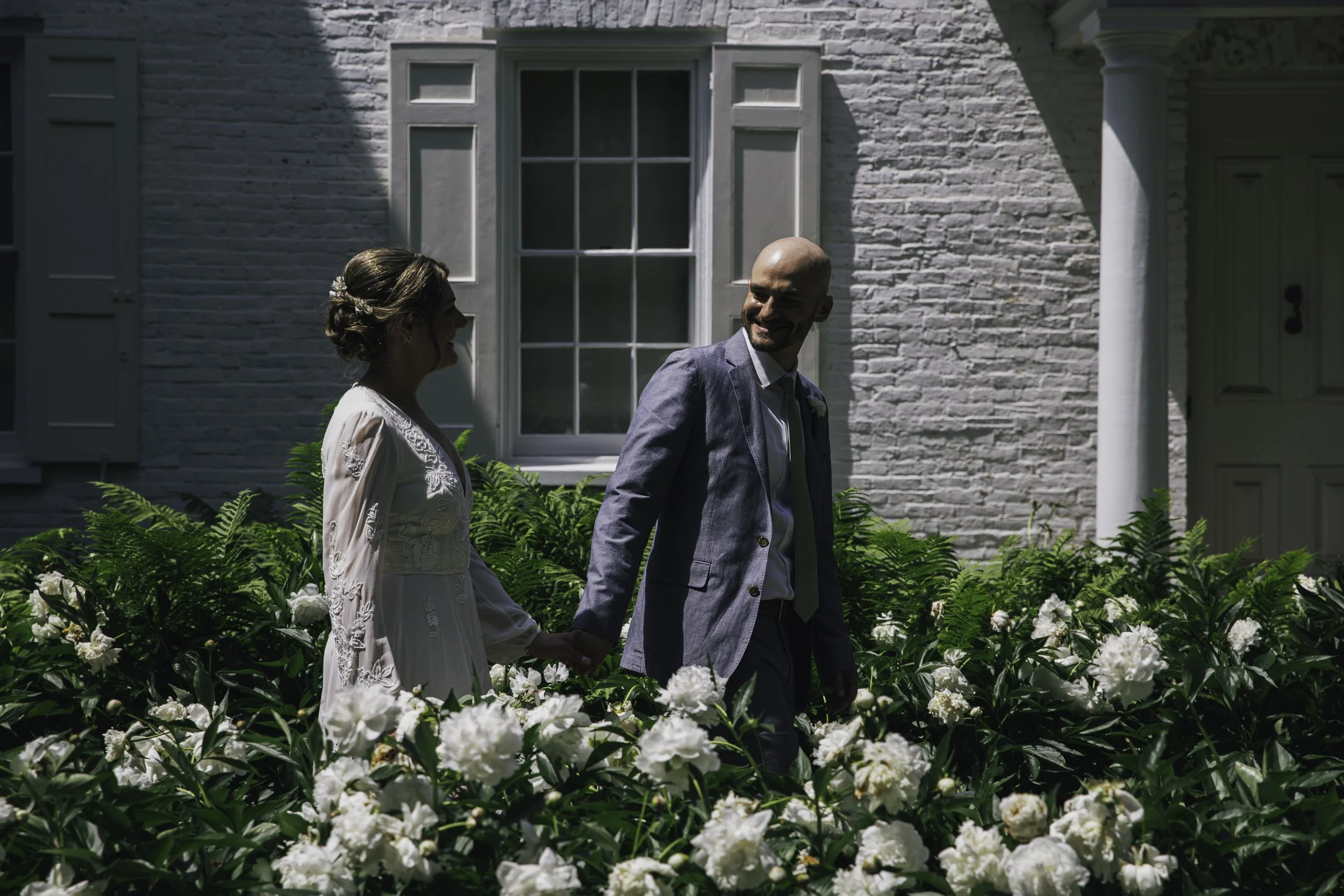 A bride and groom holding hands and walking through a garden filled with white flowers, outside a white brick house with a large window and a garage door in the background. Documentary wedding photography in NYC