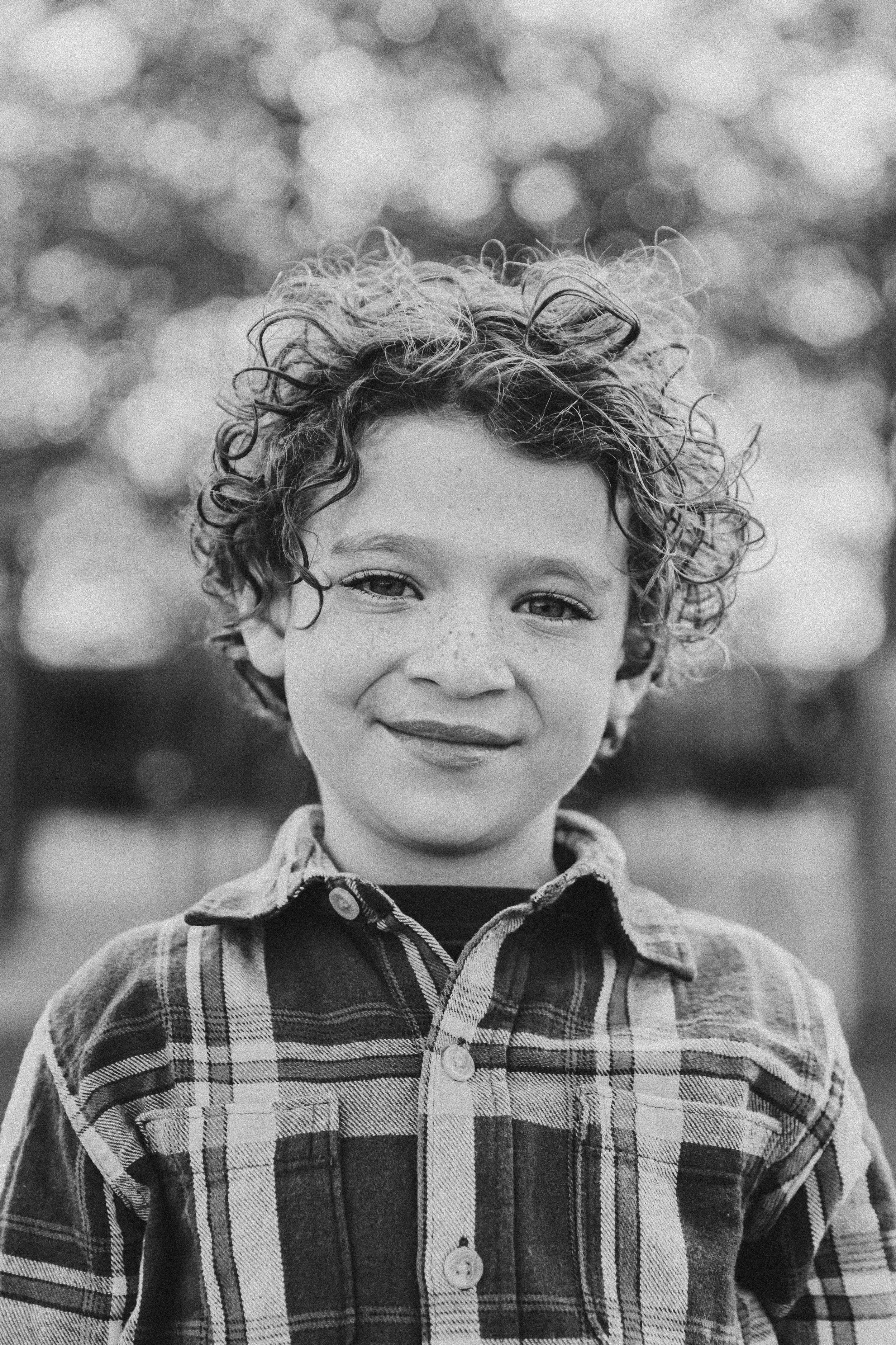A black and white photo of a young boy with curly hair, freckles, and a slight smile, wearing a plaid shirt, outdoors with blurred trees in the background. Documentary family photographer in New York City 
