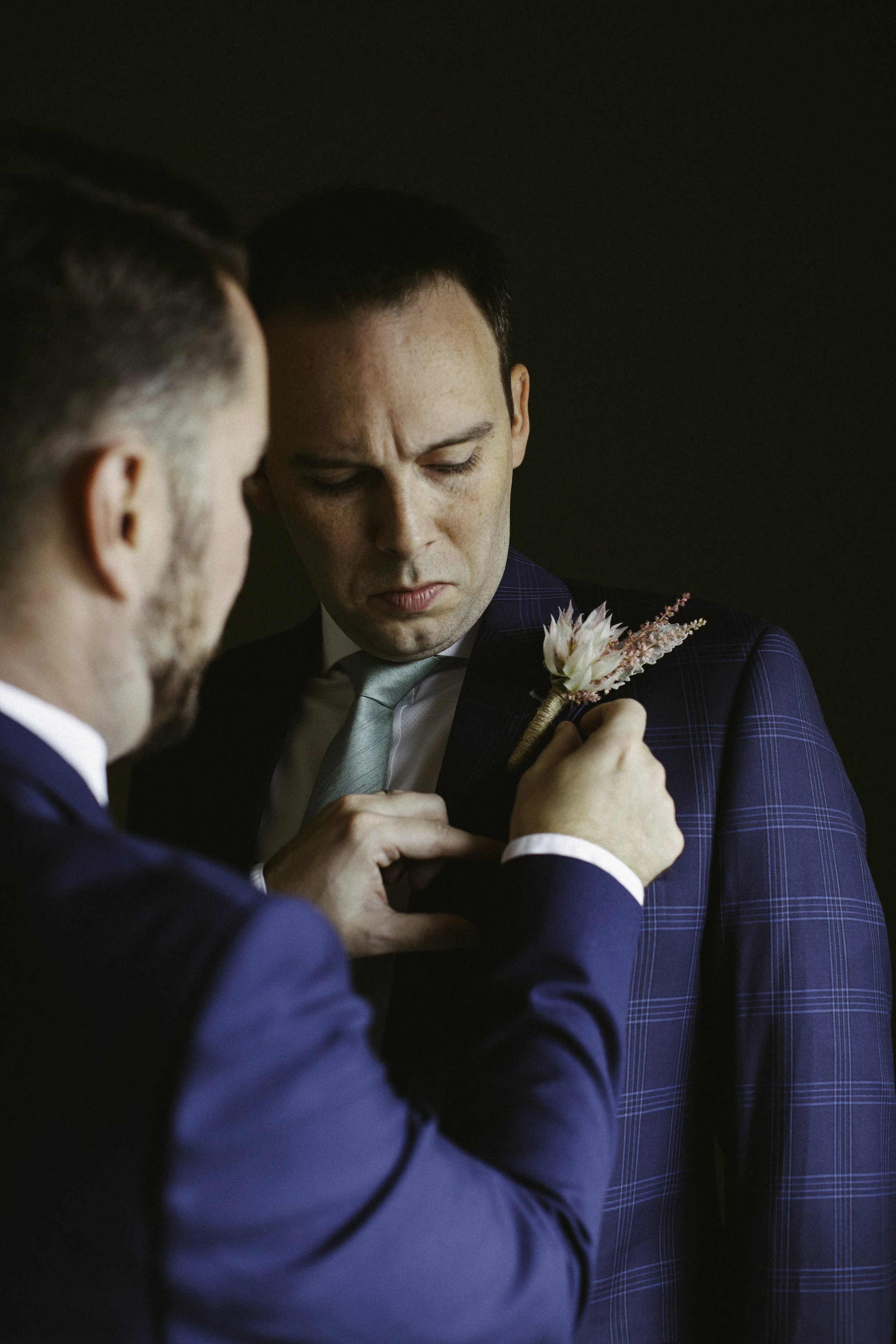 Two men in suits with boutonnières, one helping the other adjust his boutonnière on his lapel. NYC wedding photography