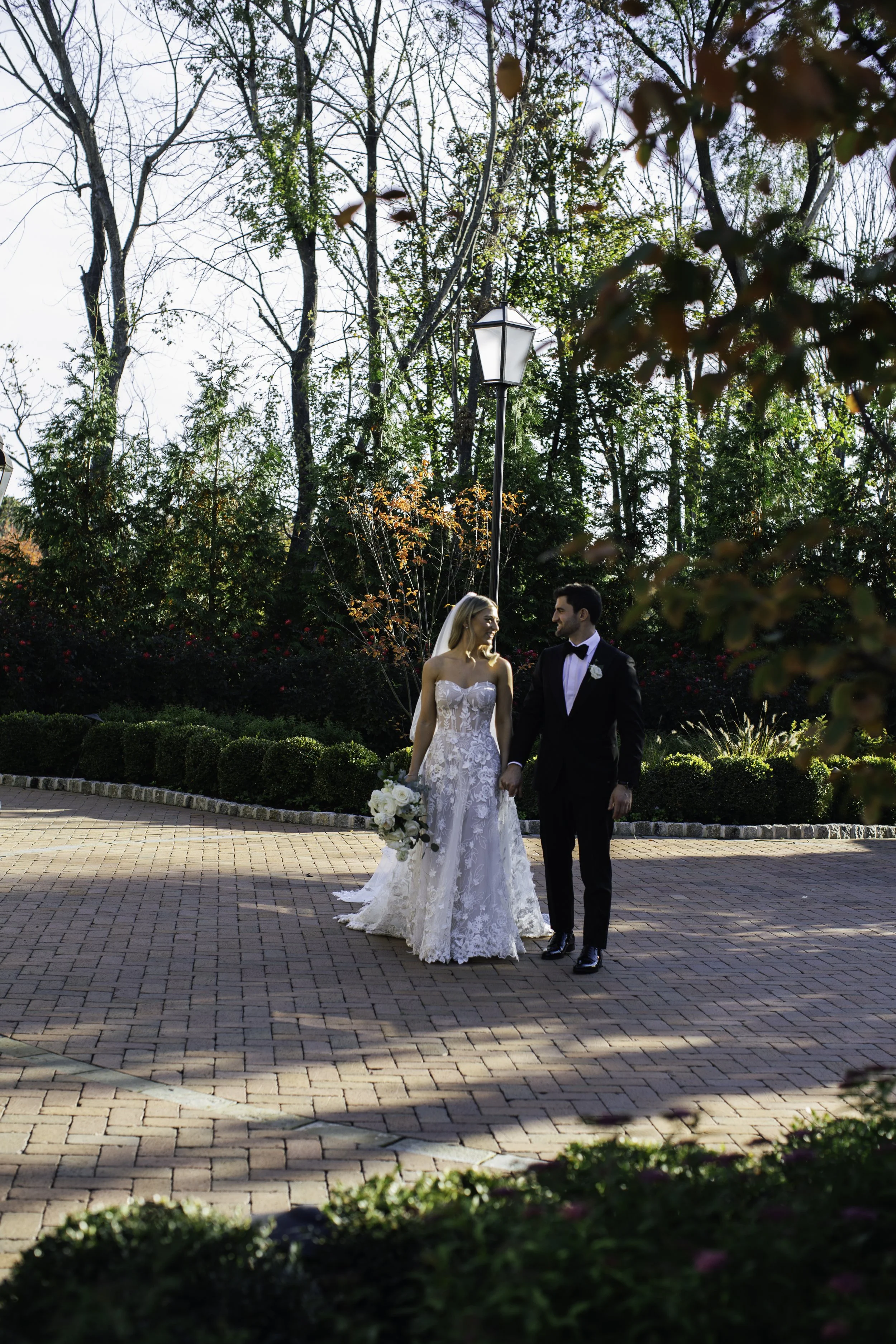 Bride and groom holding hands outdoors on a brick pathway, surrounded by trees and bushes, during a sunny day.