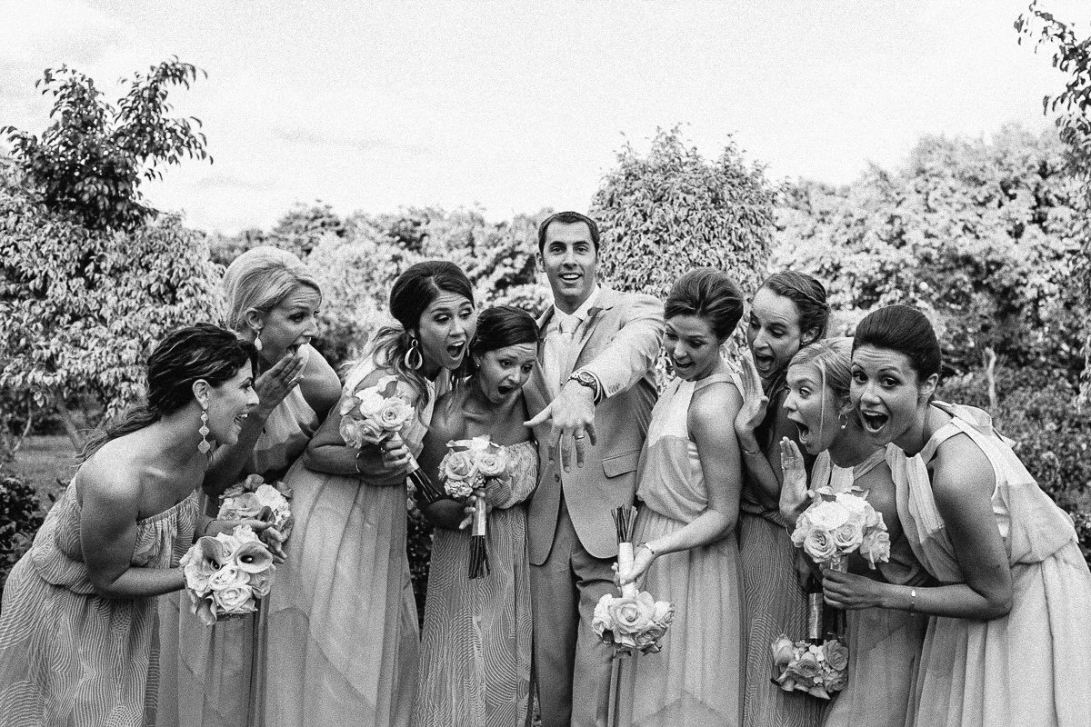 A wedding party of nine people, including the bride and groom, celebrating outdoors with surprised and joyful expressions. NYC wedding photography