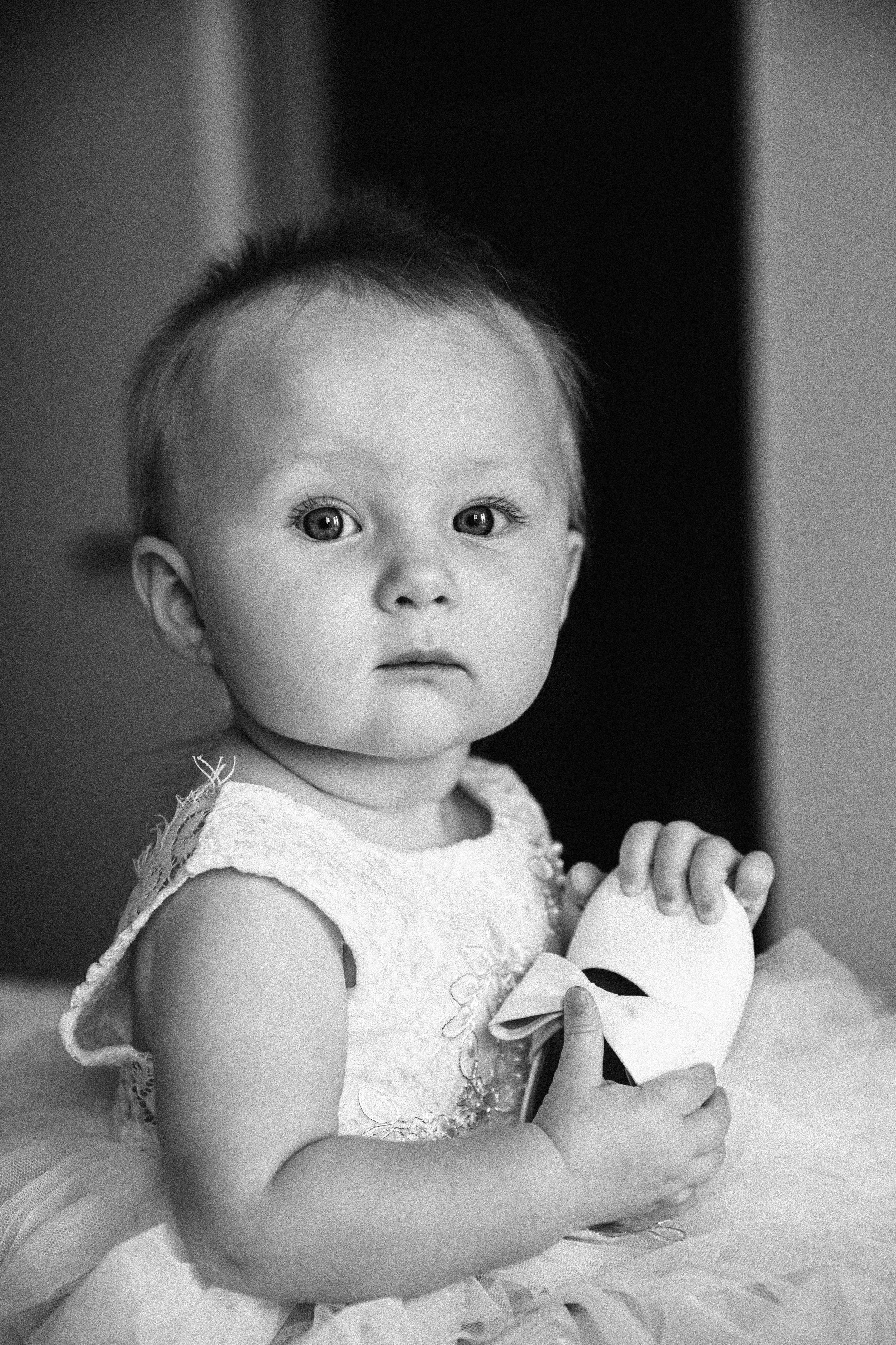 Black and white photograph of a young girl with wide eyes, wearing a sleeveless dress with lace details, holding a small item with her hands, and sitting on a soft surface.