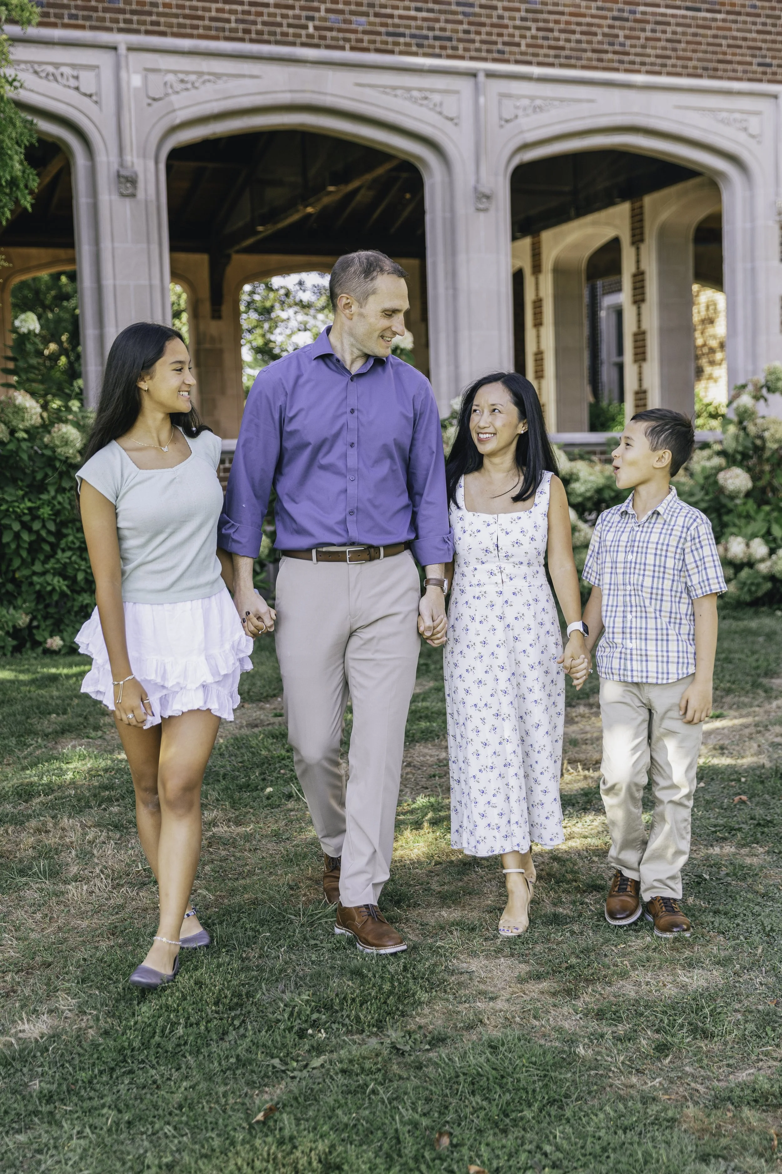 A family of five walking hand in hand outdoors in front of a large house, smiling and enjoying the sunny day.