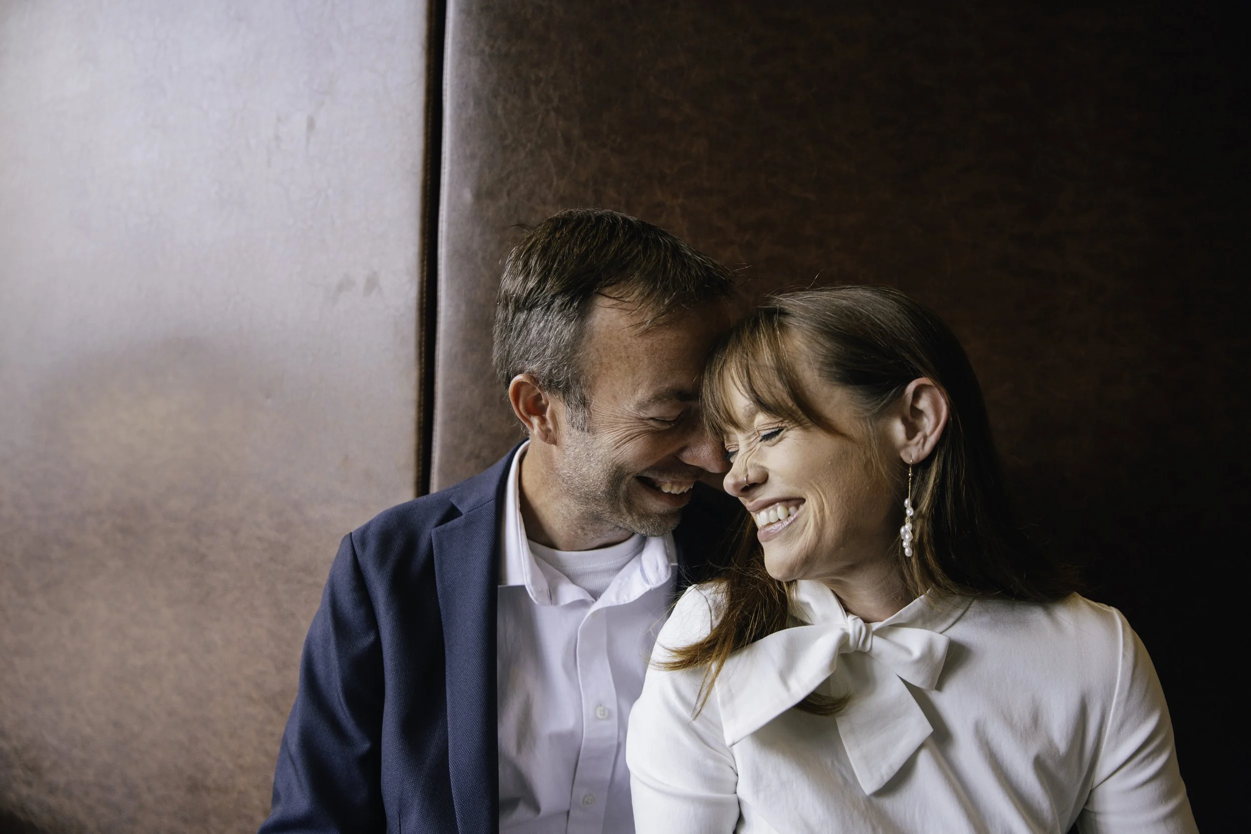 A happy middle-aged man and woman sharing a joyful close moment, smiling with their foreheads touching, seated against a brown background.