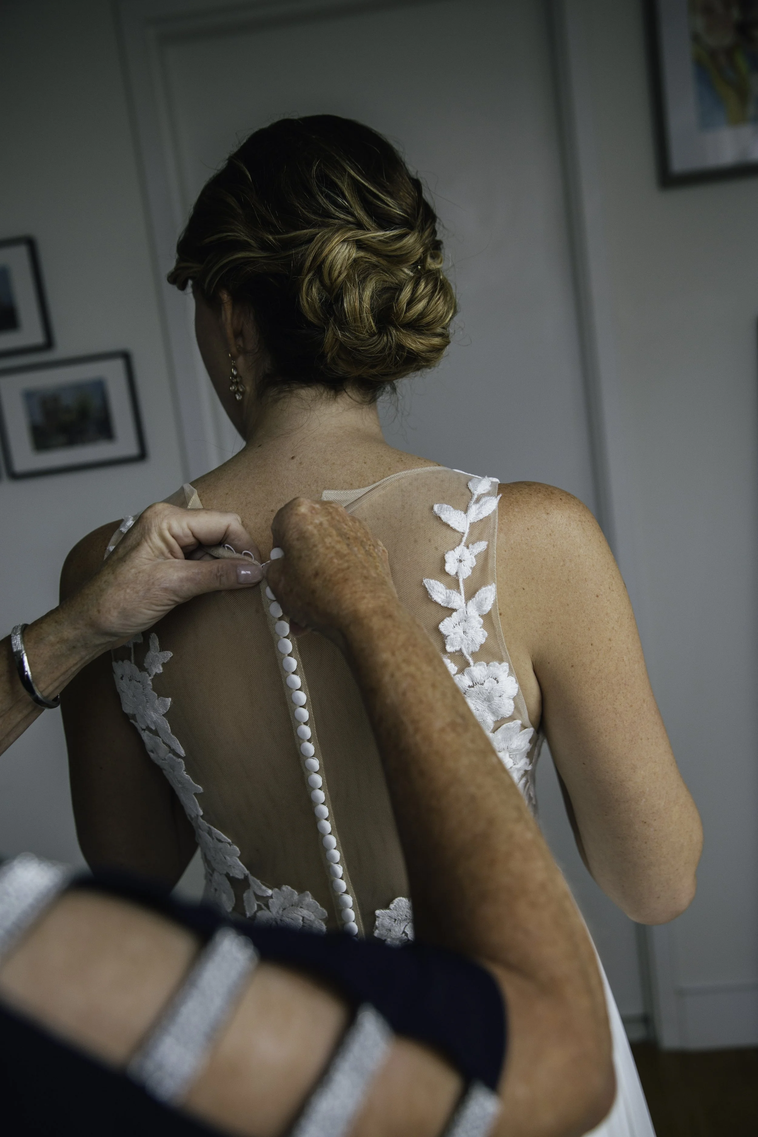 A woman is helping a bride button up the back of her wedding dress, which features floral lace details. Documentary wedding photography in NYC