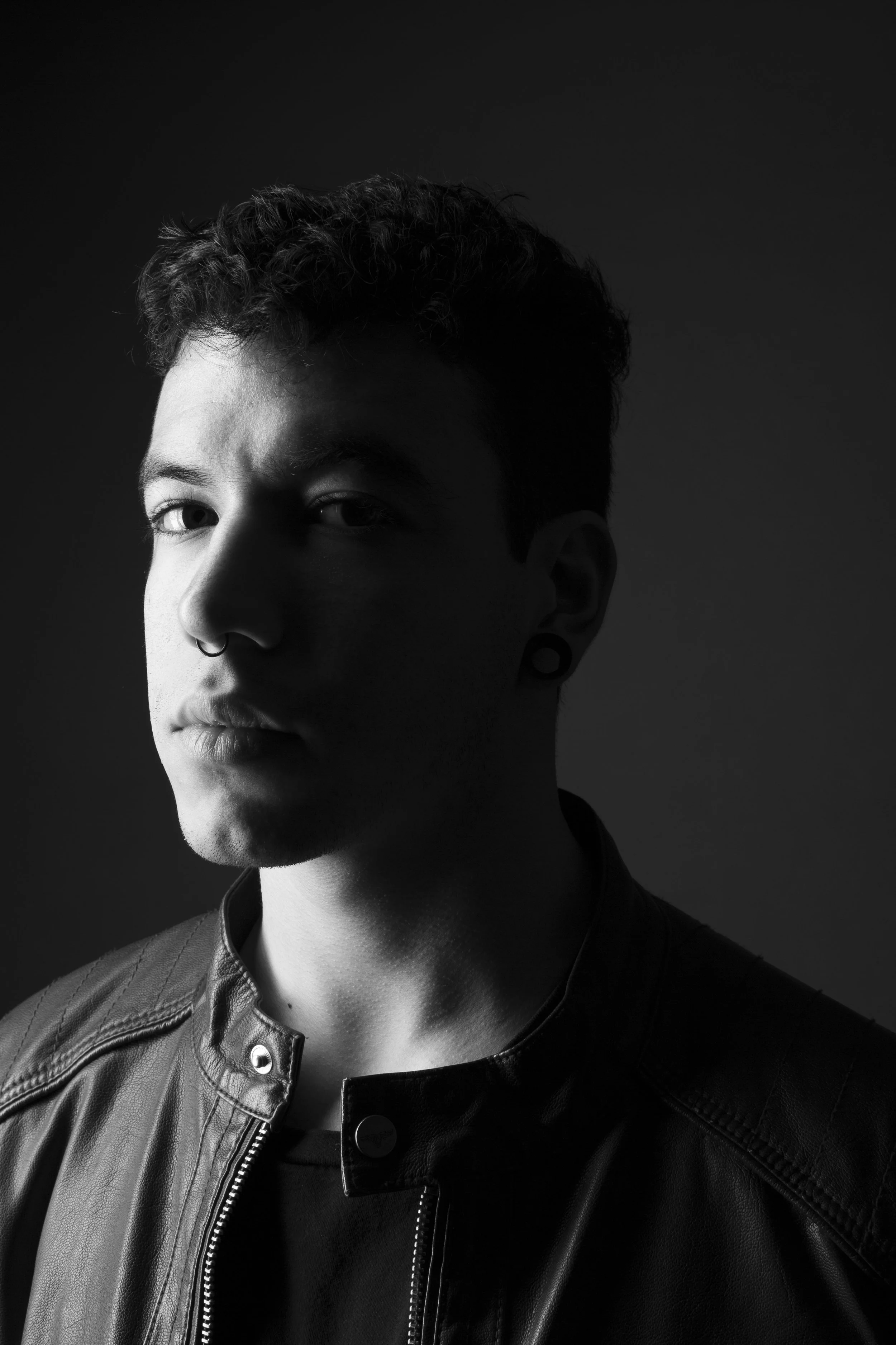 Black and white portrait of a young man with short, curly hair, wearing a leather jacket and a septum piercing, looking at the camera with a serious expression.
