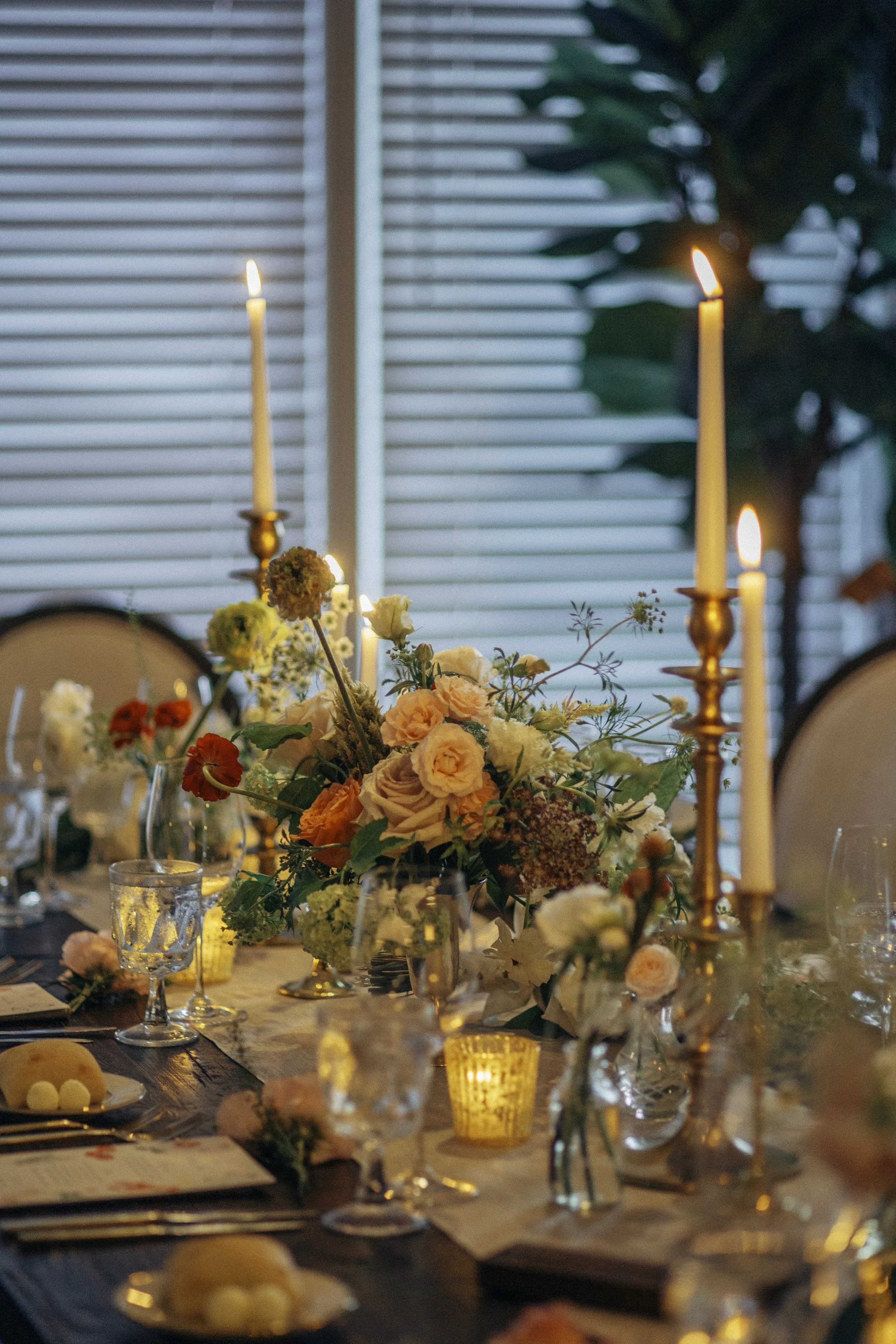 Elegant dining table with floral centerpiece, tall candleholders with lit candles, and decorative candles, set for a special occasion, in a room with large window blinds and a plant in the background.