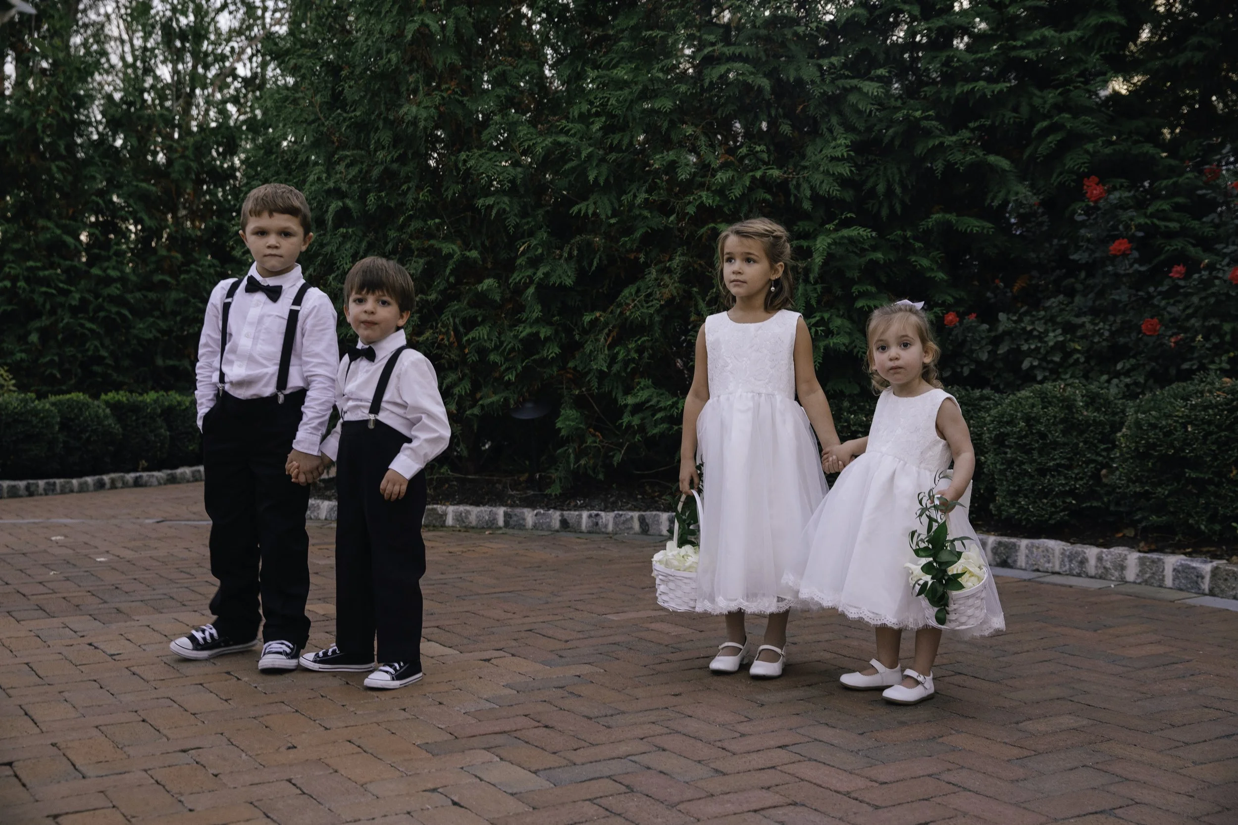 Four children in formal attire standing on a brick pathway near greenery, holding hands and baskets with flowers.