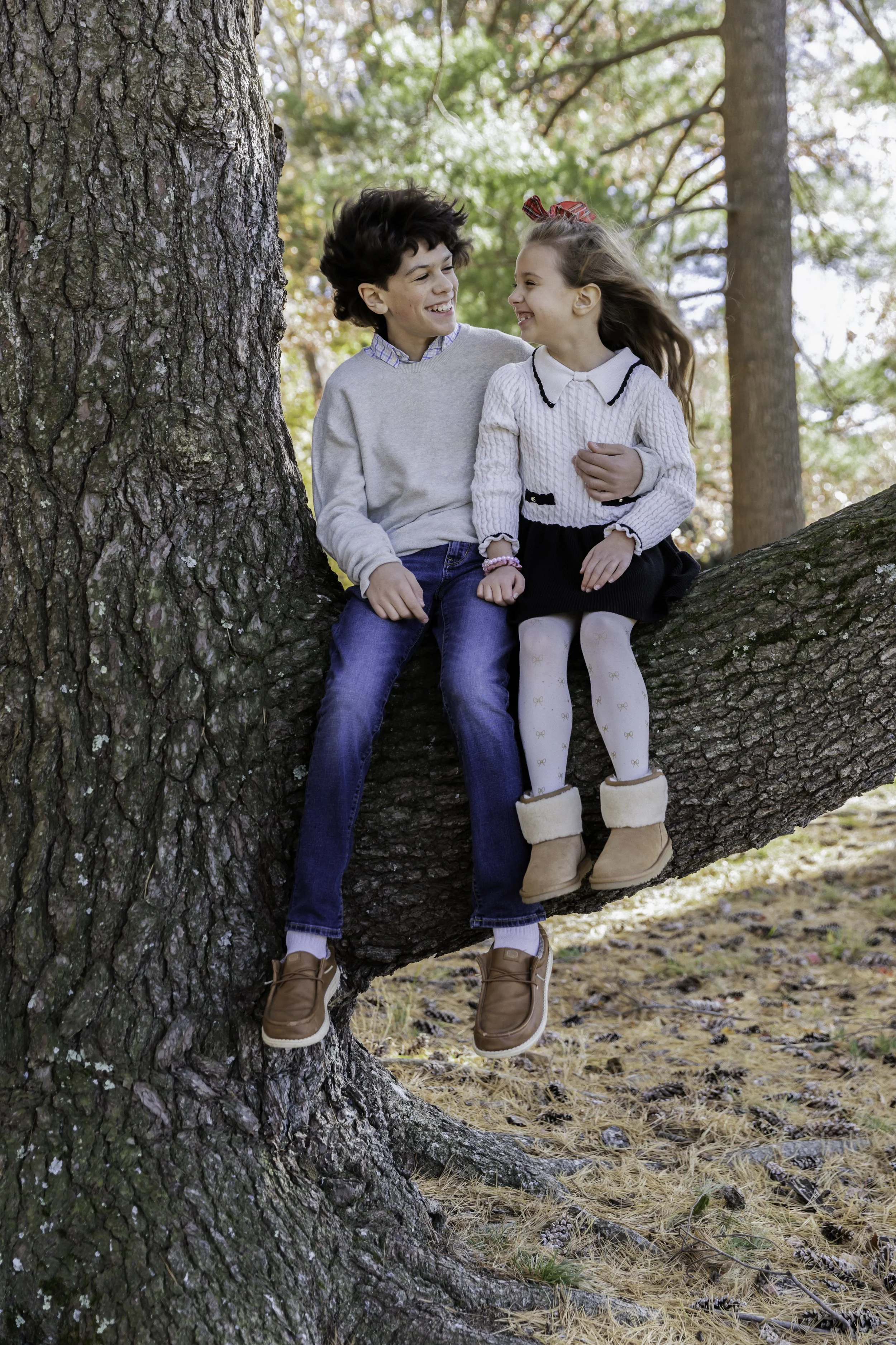 A boy and girl sitting on a tree branch, smiling and looking at each other in a forest setting during fall.
