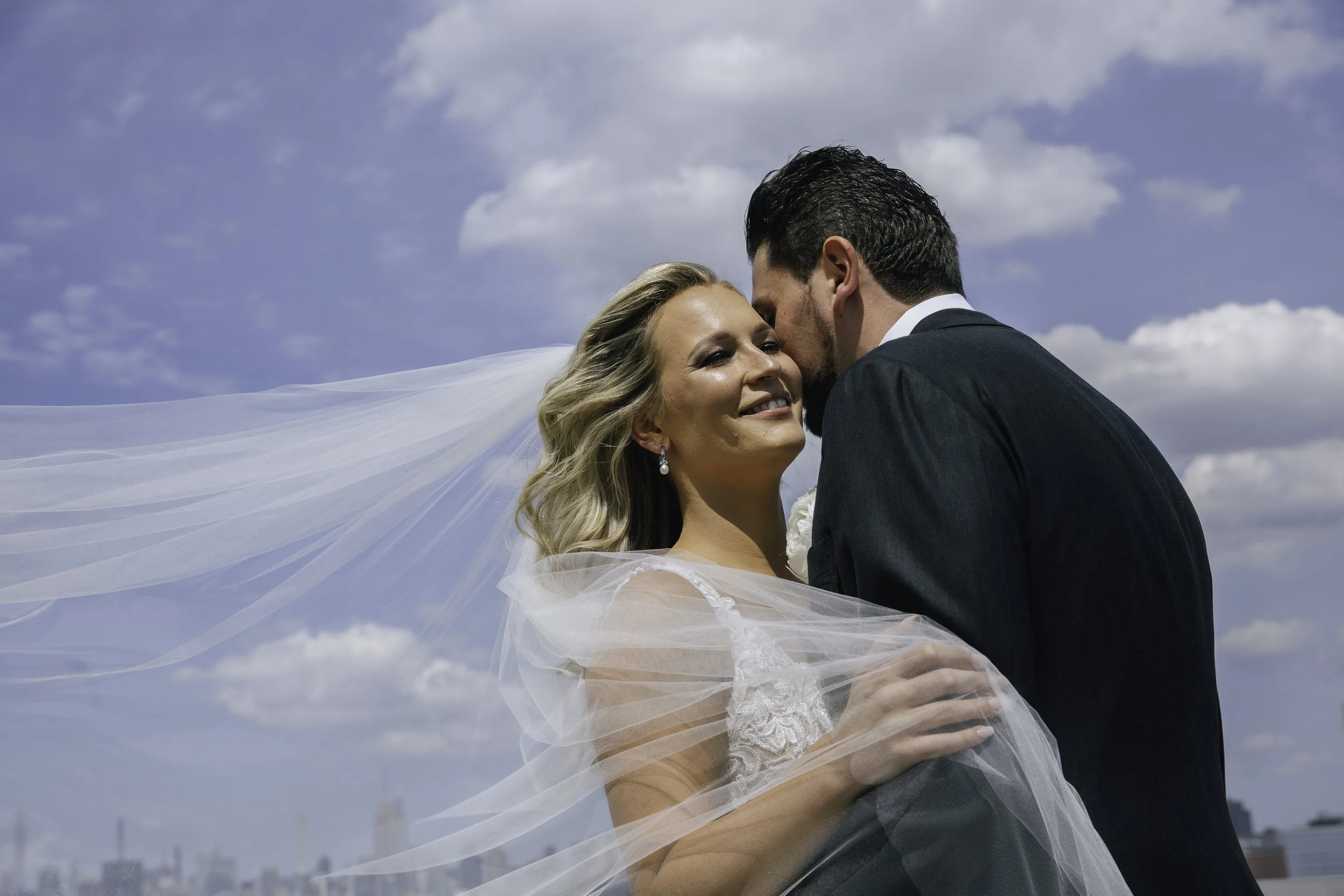 A bride and groom embrace outdoors on a sunny day with a city skyline and blue sky with clouds in the background. NYC wedding photography