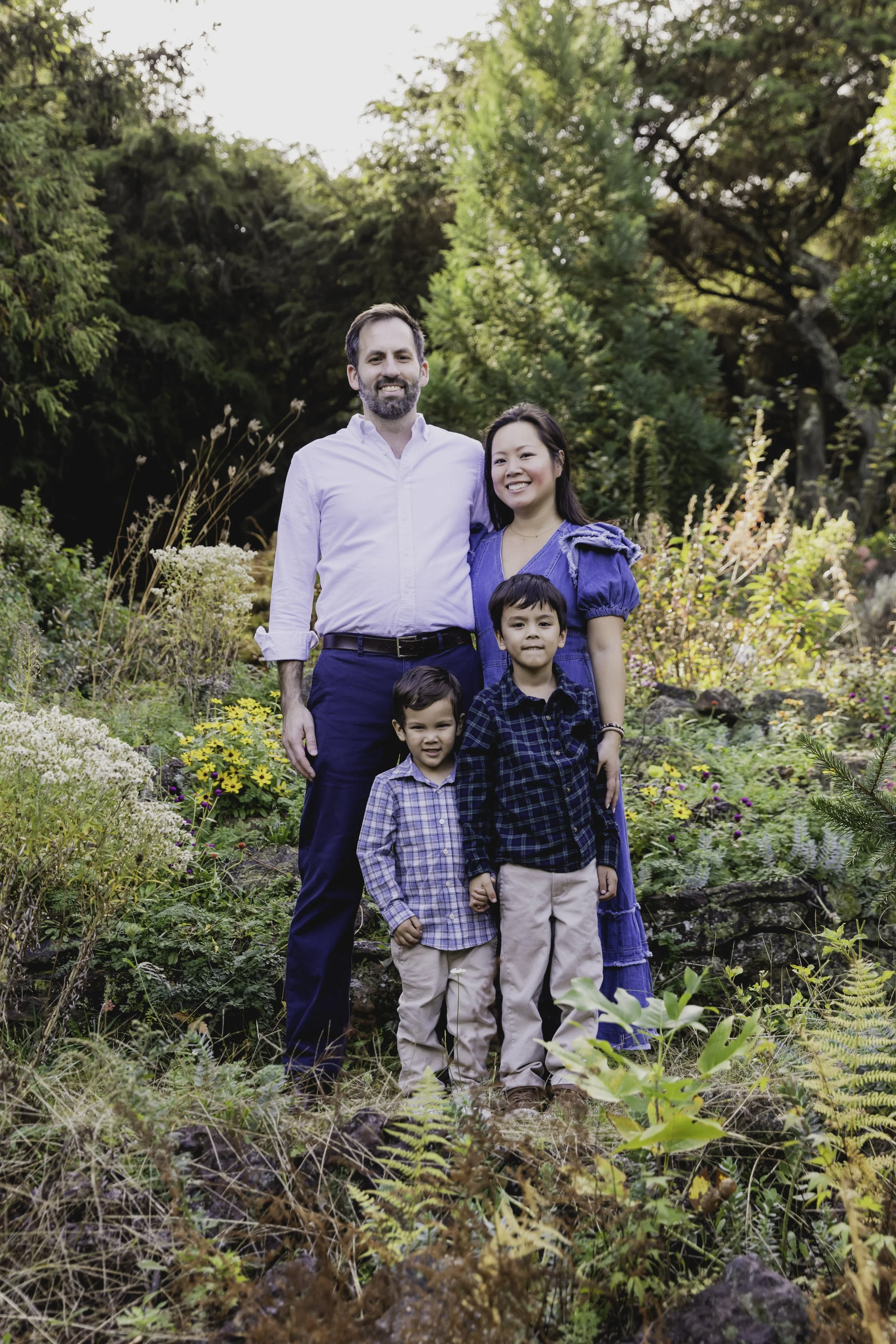 Family of four standing outdoors in a lush garden with trees and flowers, smiling at the camera. Lifestyle family photography in New York City