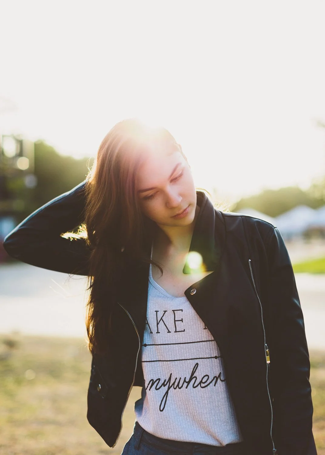 A young woman with long brown hair, wearing a black leather jacket and a white graphic t-shirt, standing outdoors during sunset, with sunlight backlighting her and creating a lens flare.