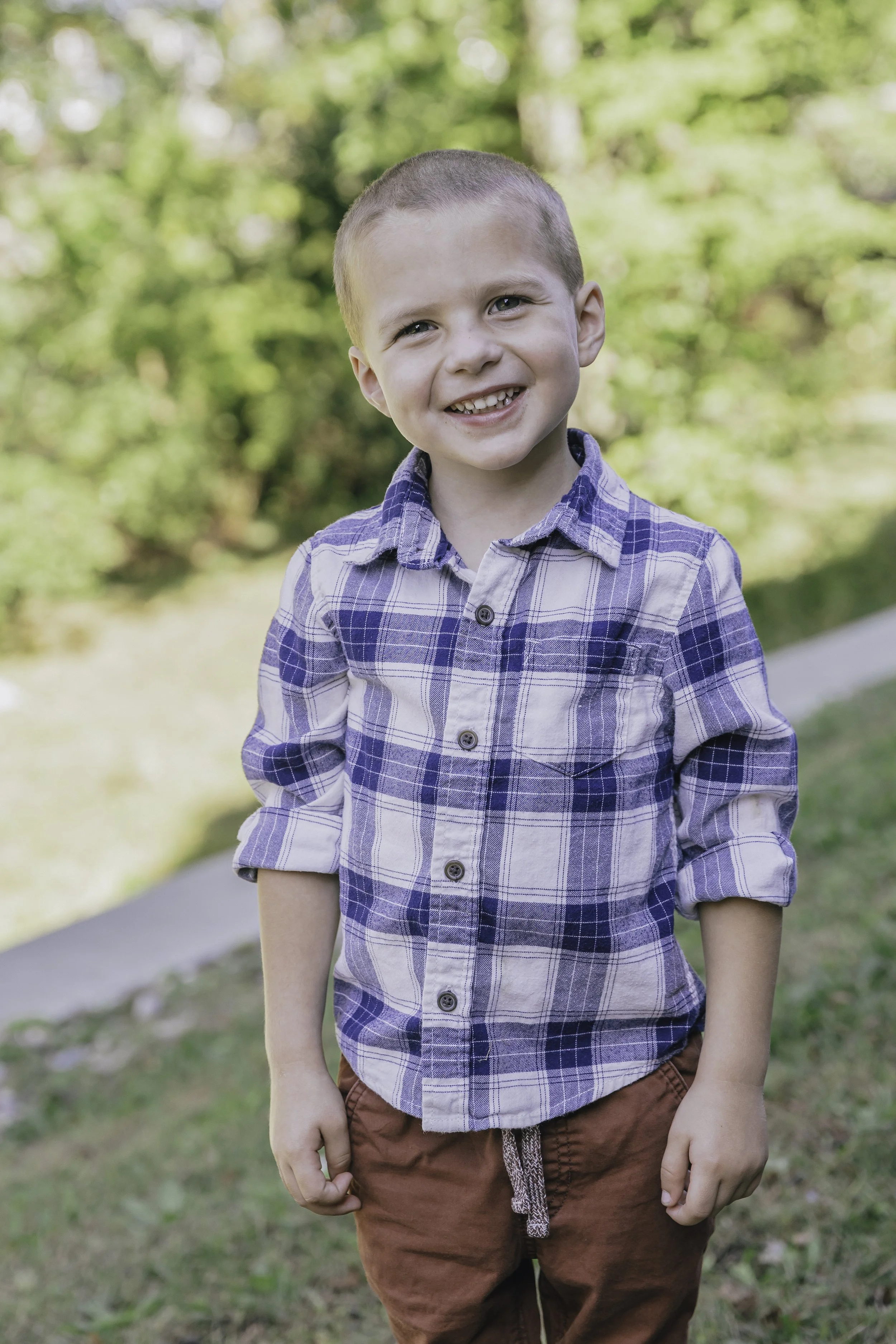 A smiling young boy with short hair wearing a blue plaid shirt and brown pants, standing outdoors in a park with green trees and grass in the background.