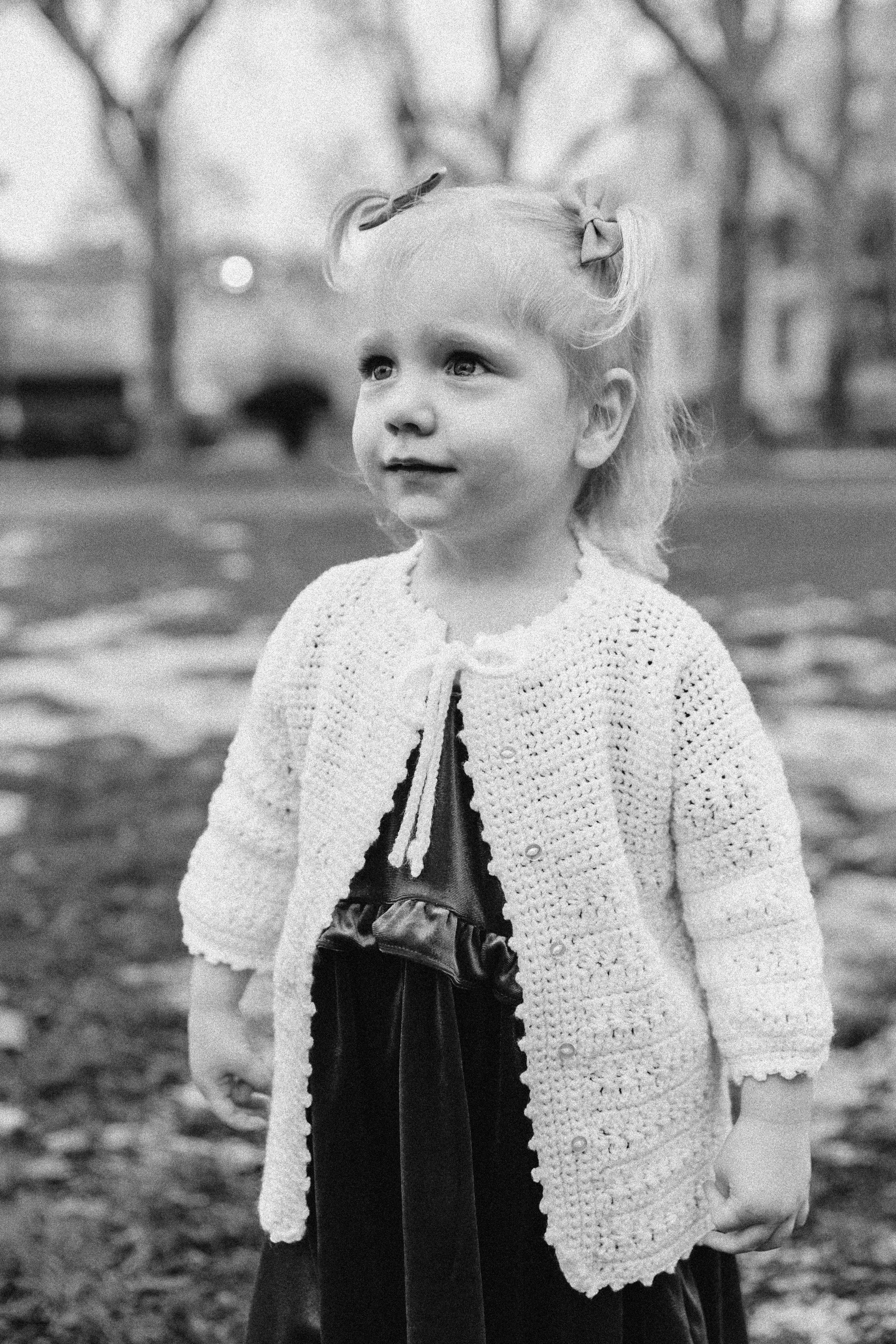 A young girl with light curly hair wearing a knitted cardigan standing outdoors in a park with trees in the background.