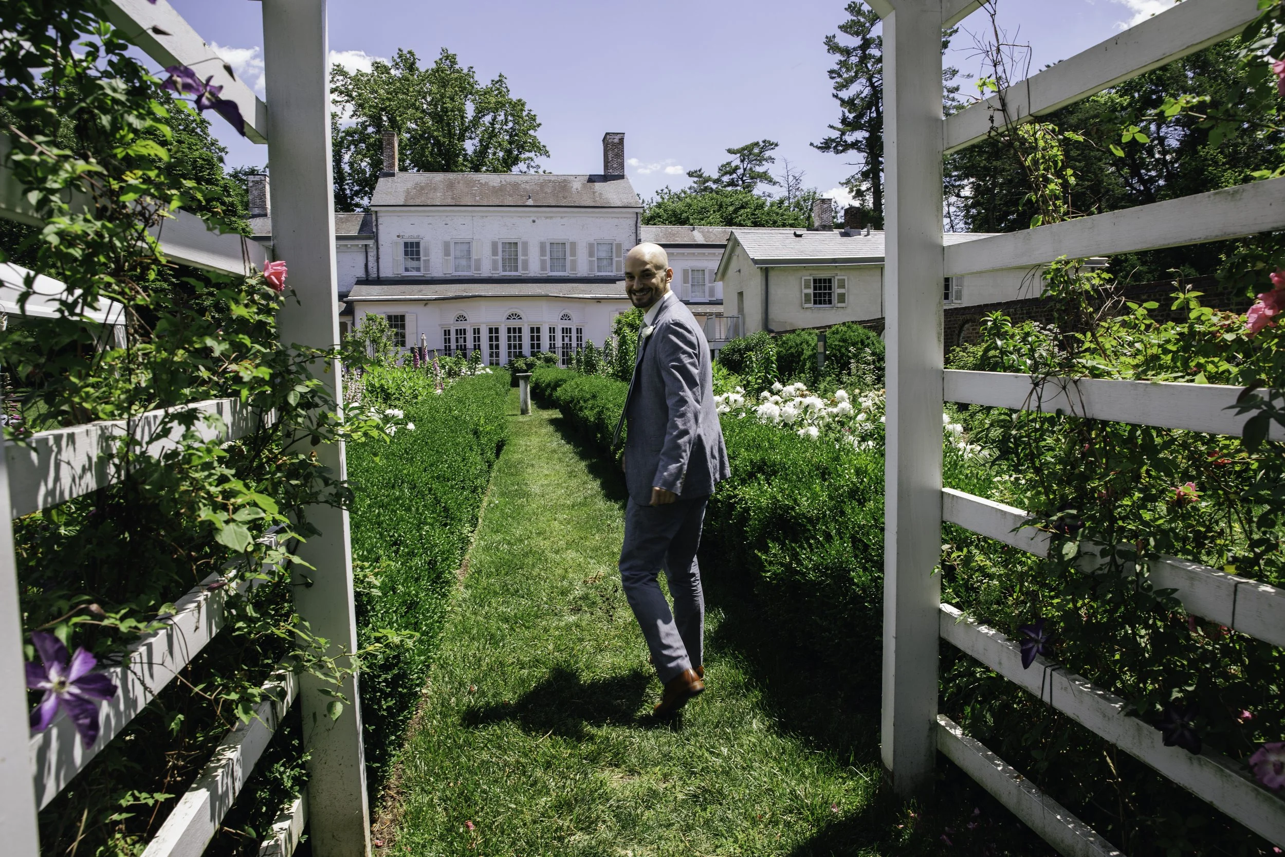 A man in a gray suit stands in a garden pathway surrounded by lush greenery and flowers, Documentary wedding photography in NYC